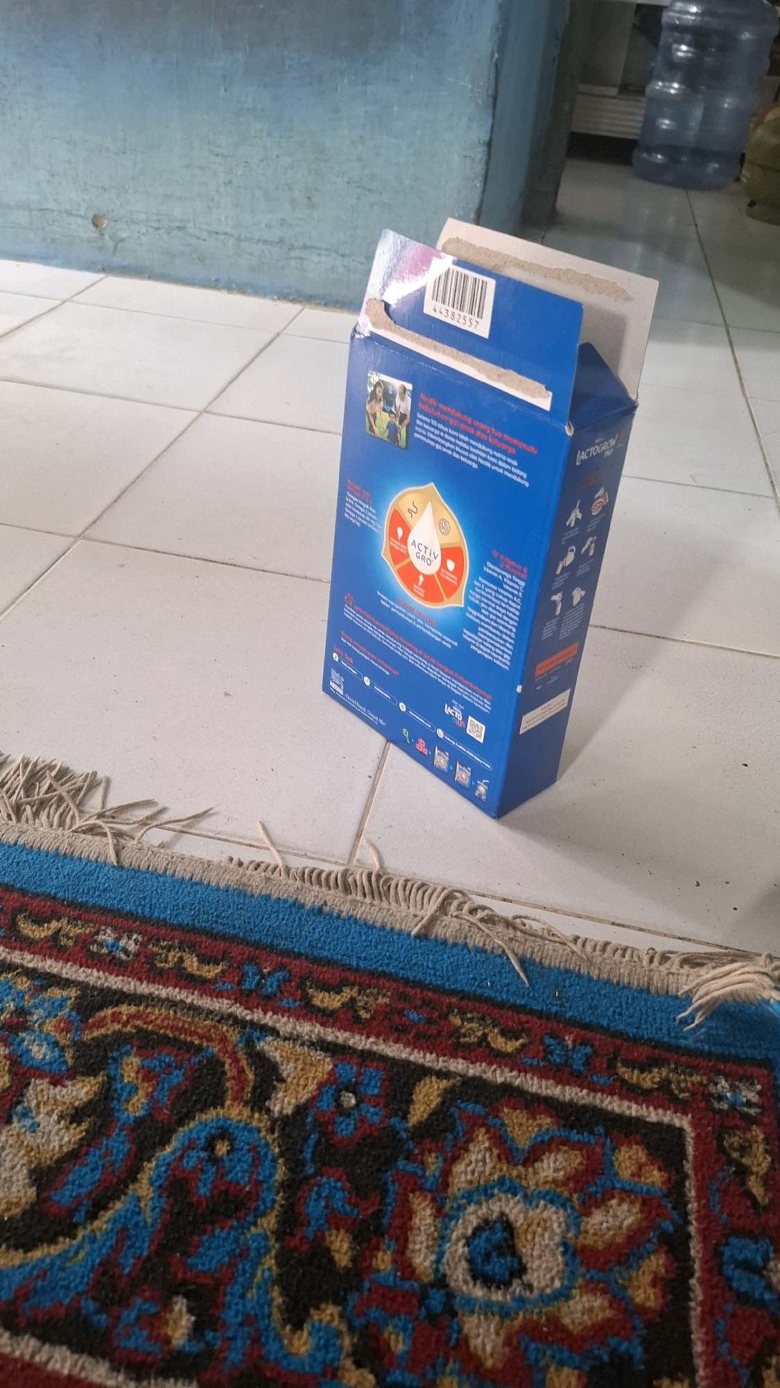 A close-up, slightly angled shot shows an open blue cardboard box of Lactogrow Activ-Gro Pro milk powder on a white tiled floor. The box is positioned near a blue rug with intricate patterns in red, yellow, and black. The lid of the box is partially open, revealing the inside. The background shows a light blue wall and a large clear plastic water bottle. A barcode is visible on the box, and text in Indonesian is printed on its sides. The scene appears to be indoors, possibly a residential or commercial space in Bandar Lampung, Indonesia.