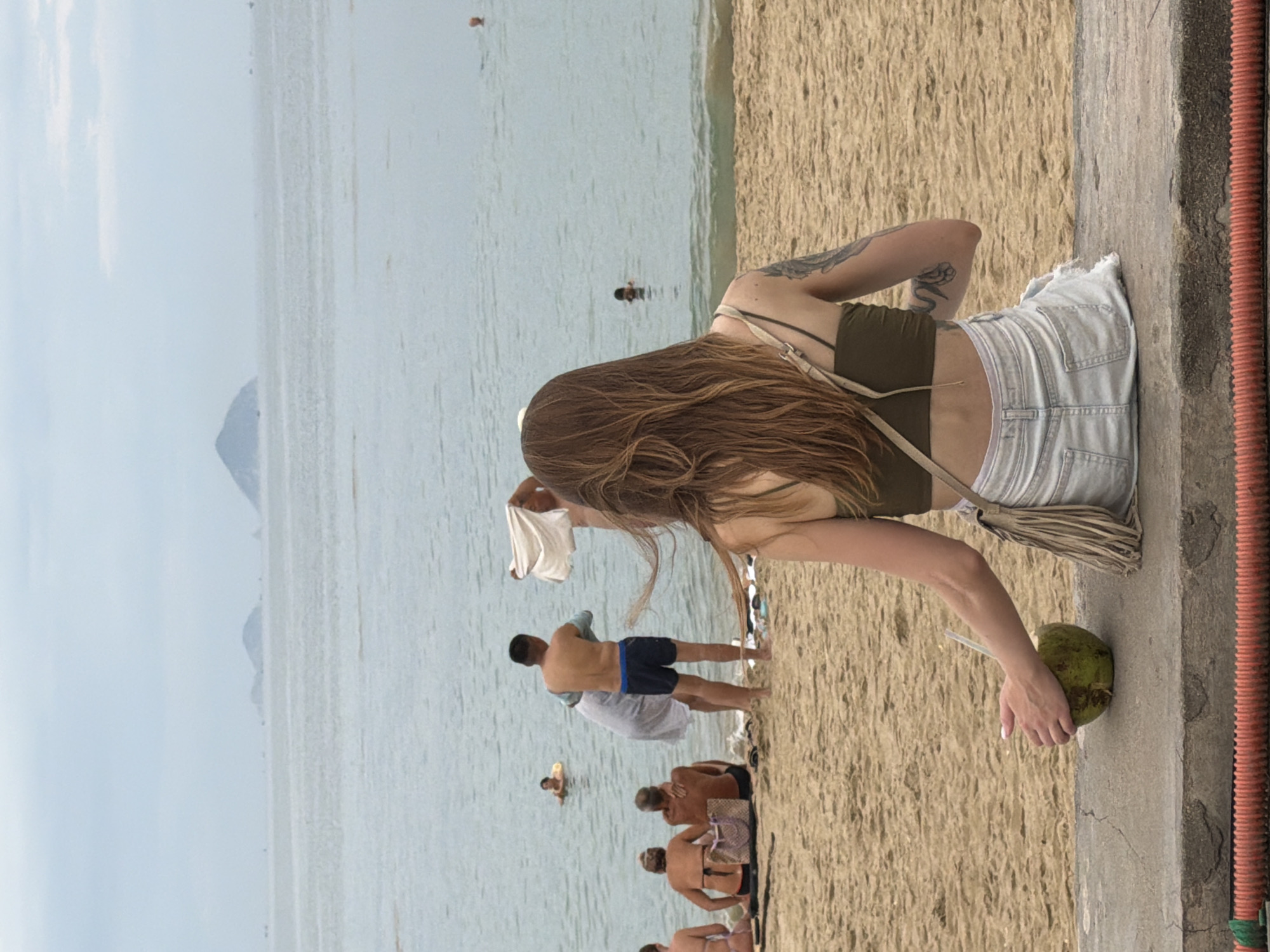 A medium shot captures a woman from behind, seated on a concrete ledge overlooking a sandy beach and the ocean. The woman has long, wavy brown hair and is wearing a green camisole and ripped denim shorts. Her left arm is resting on her lap, holding a halved green coconut with a straw. A light-colored bag with fringe hangs from her shoulder. In the background, several people are on the beach and in the shallow water. Some are swimming or standing, while others are seated on the sand. Further out in the ocean, a few small islands are visible on the horizon under a cloudy sky. The scene appears to be a typical day at the beach in Nha Trang, Vietnam, with a relaxed atmosphere.