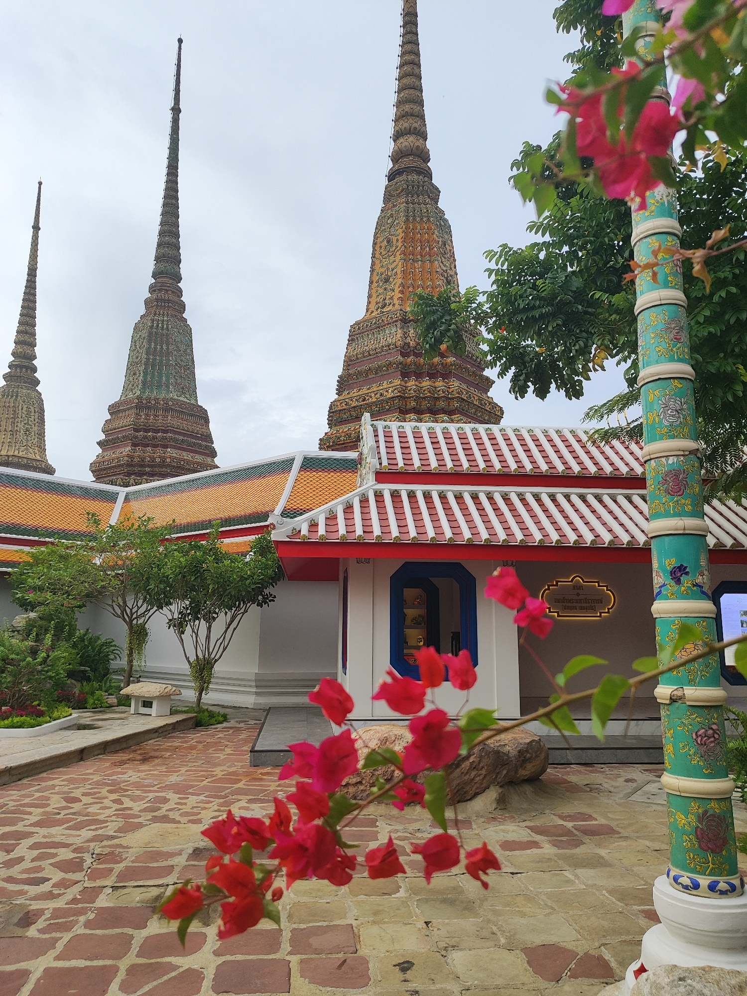 The image depicts a section of Wat Pho, also known as the Temple of the Reclining Buddha, located in the Phra Nakhon district of Bangkok, Thailand. In the foreground, vibrant pink bougainvillea flowers are in soft focus, partially obscuring a decorative teal-colored pillar adorned with floral motifs. Behind the pillar, a low-slung building with a red and white tiled roof is visible. The building features white walls and a blue-trimmed entrance. To the left of the building, several ornate, cone-shaped stupas rise against a cloudy sky, characteristic of Thai temple architecture. The ground is paved with reddish-brown cobblestones. A small, bonsai-like tree and lush green foliage are scattered throughout the scene, adding to the serene temple environment. The overall impression is one of tranquil beauty and intricate cultural design.