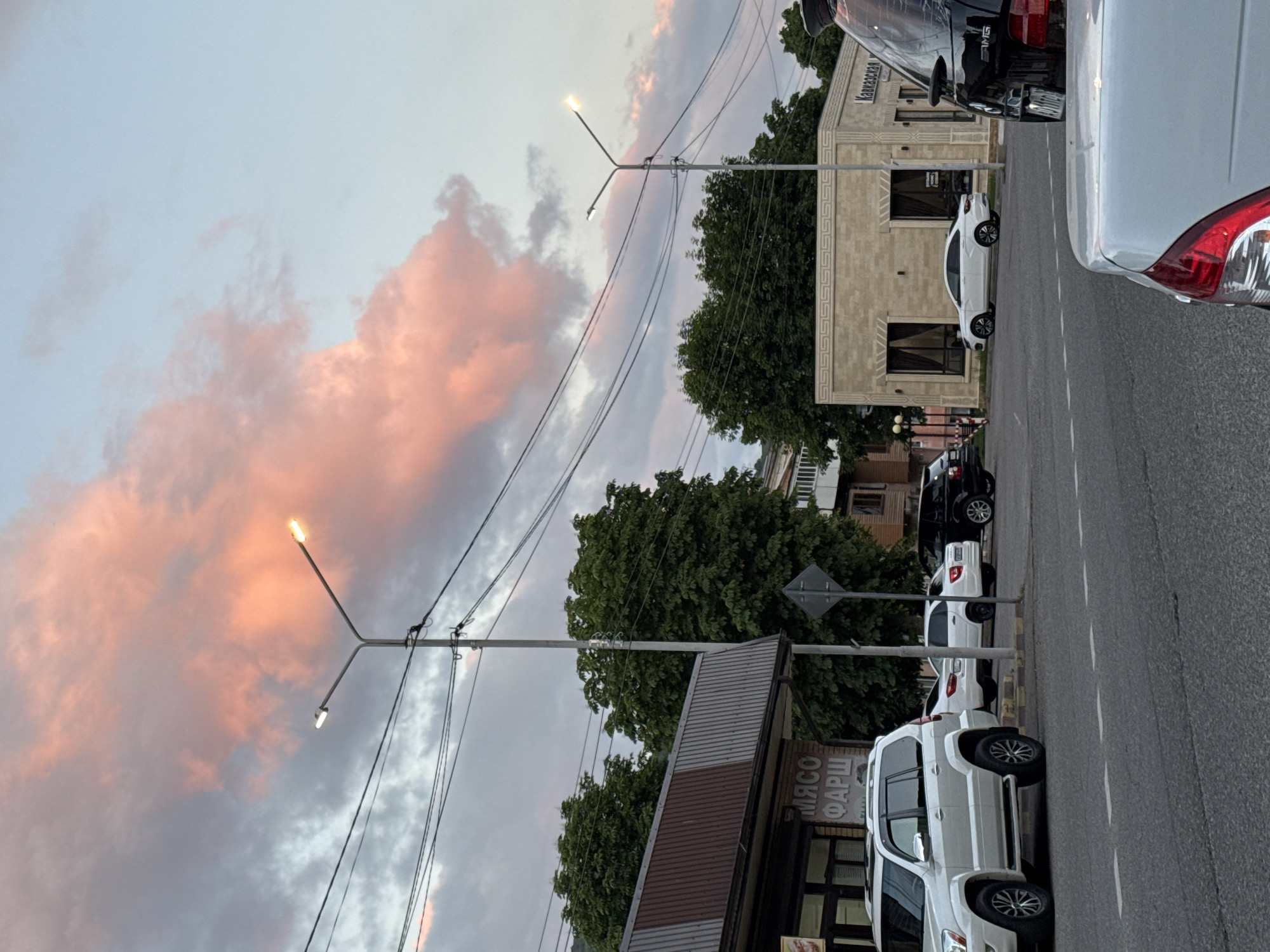 The image captures a street scene in Karachayevsk, Russia, during the golden hour. A large, pink-tinged cloud dominates the sky, casting a warm glow. A street lamp with two illuminated bulbs stands prominently in the foreground, surrounded by power lines. To the left, a building with a metal awning displays Russian text that reads "МЯСО ФАРШ" (Meat Mince). Several cars are parked along the street, including a white SUV, two white sedans, and a black SUV. Across the street, a modern, beige building with ornate trim and dark windows is visible, with Russian text reading "Кавказская" (Caucasian) above one of the windows. Trees line the street, adding a touch of greenery to the urban landscape. The road in the foreground shows dashed white lines, indicating a marked lane. The overall atmosphere is tranquil, with the soft lighting of the sunset creating a peaceful ambiance.