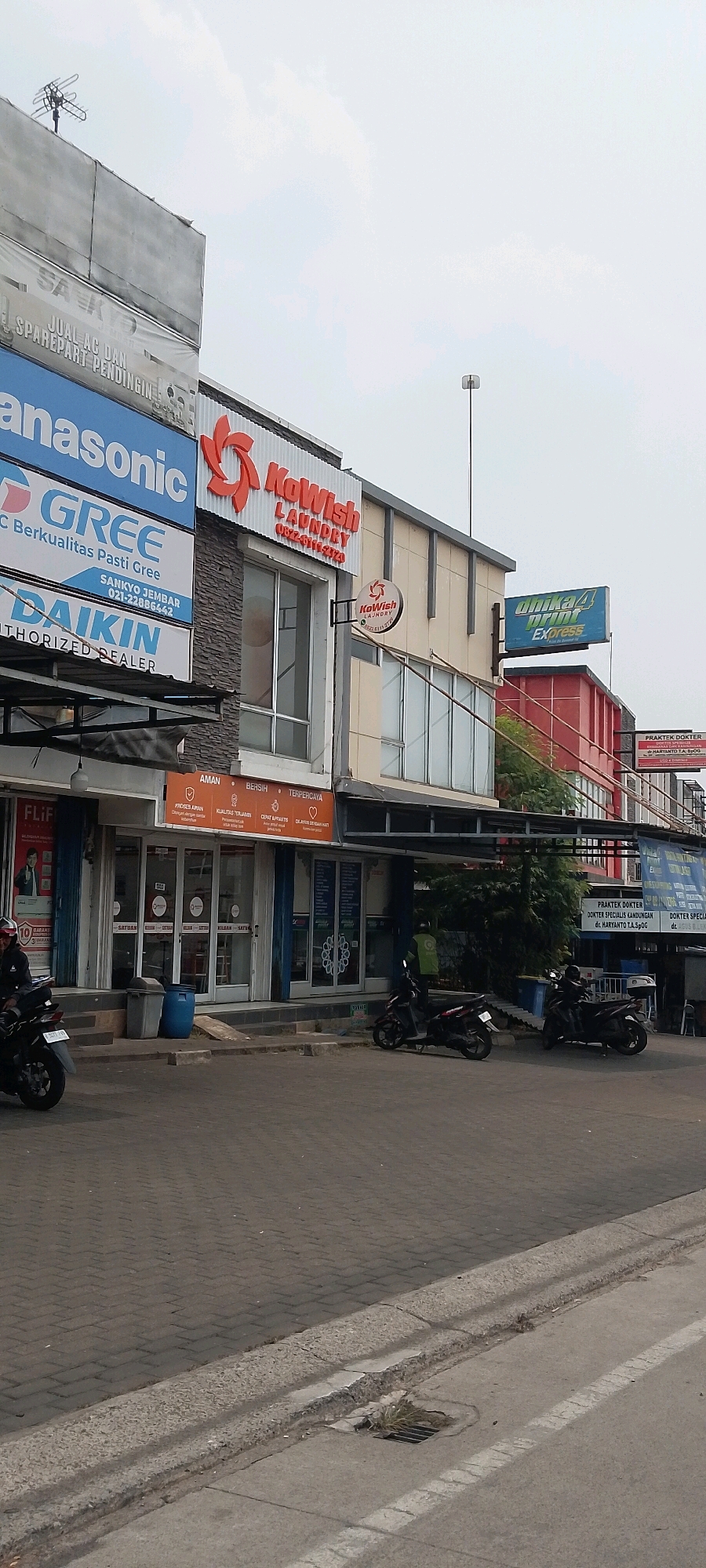The image shows a street scene in Cileungsi, Indonesia, with a row of commercial establishments. In the foreground, a paved sidewalk and road are visible, with some motorcycles parked and one rider on a motorcycle in the left portion of the image. Behind the sidewalk are buildings with various signs.
The building on the left features prominent signage for "Panasonic," "GREE," and "DAIKIN," indicating electronics and appliance sales. To the right of these, a "KoWish LAUNDRY" sign is displayed prominently. Further to the right, another "KoWish LAUNDRY" sign is visible. A larger blue sign on a higher building reads "Unika Print Express." There are also smaller signs indicating "PRAKTEK DOKTER" (Doctor's Practice).
The buildings appear to be multi-story, with glass storefronts and windows. Some of the establishments have awnings or covered walkways. The sky is overcast. The environment suggests a commercial district within a populated area.