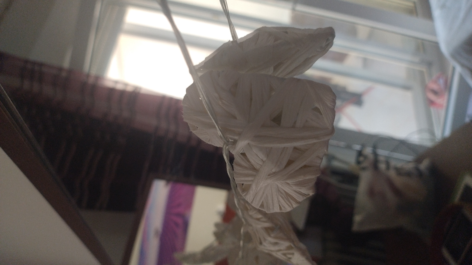 The image shows a close-up of a decorative star made of woven white material, possibly paper or straw, suspended by thin wires. The star appears to be part of a garland or string lights, with other similar star shapes visible to the left and right. The background is blurred, but suggests an indoor setting with a window showing daylight and a glimpse of a dark curtain with a striped pattern. There is also a reflection in a surface to the left, possibly a mirror or a glass table, showing a pinkish object. The scene is lit by natural light coming from the window. The image does not contain any information that would allow for its placement in Perea, Greece.
