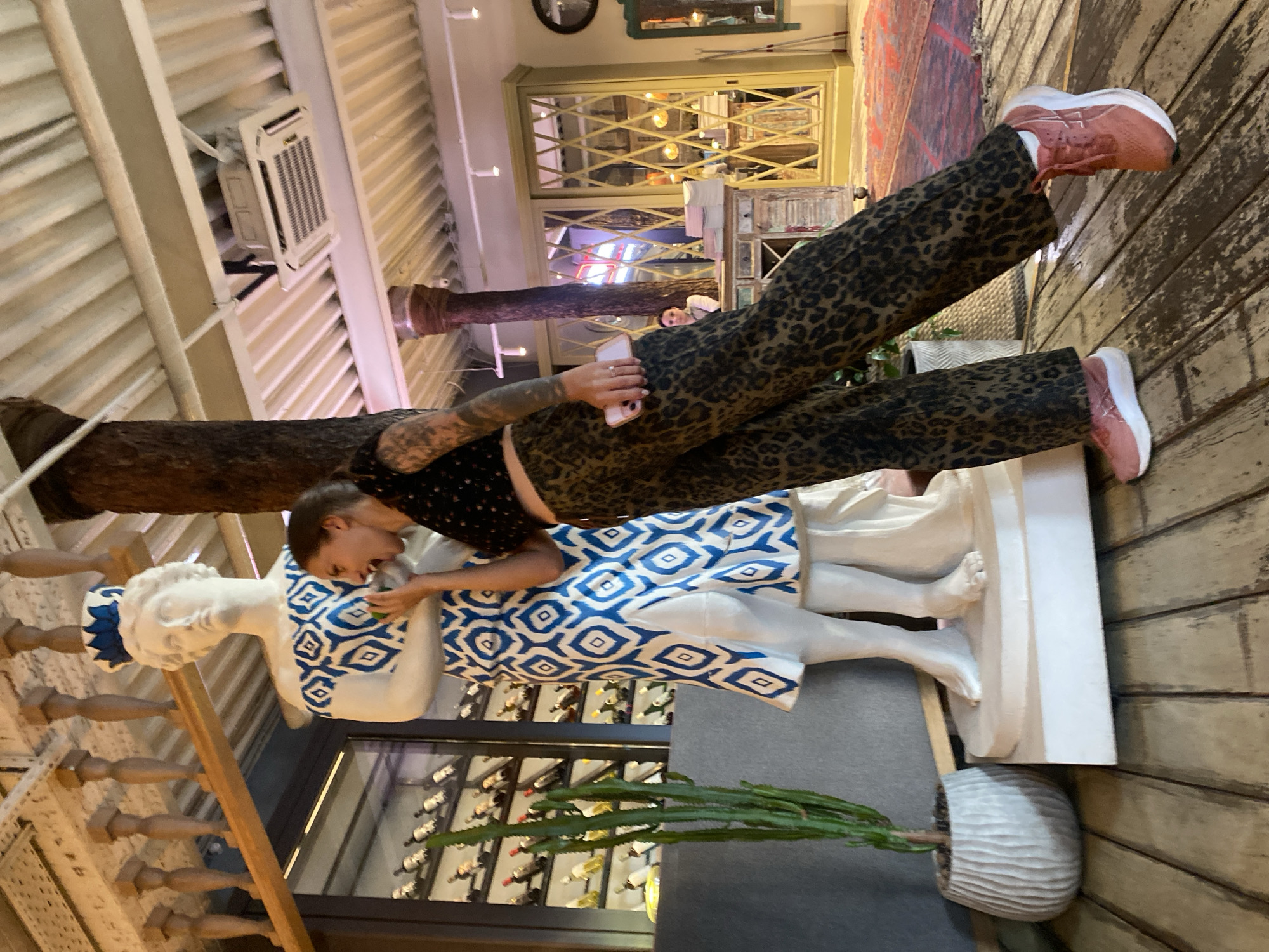 The image shows a woman in a leopard print pantsuit and a dark patterned crop top, playfully interacting with a white statue of a woman wearing a blue and white patterned dress. The statue is tall and stands on a white pedestal. The woman is leaning towards the statue, with her mouth open as if speaking to it or taking a bite, and holding a small object, possibly a piece of fruit, near its face. The scene appears to be indoors, with wooden floorboards and a ceiling with exposed beams and lighting fixtures. In the background, there are shelves stocked with wine bottles, a large cactus in a decorative pot, and some ornate furniture. The location is identified as Solnechnoye, Russia.