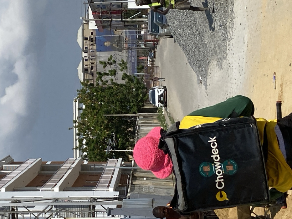 The image depicts an outdoor urban scene in Ikate, Nigeria, under a partly cloudy sky with prominent dark grey clouds. In the immediate foreground, a person is seen from behind, wearing a bright pink fluffy bucket hat, a yellow and green long-sleeved shirt, and black pants. They are carrying a large black delivery bag emblazoned with the "Chowdeck" logo in white and yellow. Bicycle handlebars are visible at the bottom of the frame, indicating the person's mode of transport. To the left of this central figure, a second individual, partially visible, stands beside a wheelbarrow filled with sand. To the right, a third person, a male in a high-visibility yellow-green vest and dark shorts, stands next to a large, conical pile of grey gravel, holding a tool (likely a shovel).
The midground features an unpaved dirt and gravel road extending into the distance. A white Lexus SUV is positioned on this road, either parked or moving slowly. Further down the road, a lone pedestrian walks away from the camera towards a distant gate or barrier. On the left, a grey concrete wall runs parallel to the road, bordering a property. Behind this wall, a multi-story building with white exterior walls, brown sections, and white-railed balconies suggests new construction or recent development. Green foliage and trees are visible beyond the wall. On the right side of the road, several dark-colored vehicles are parked, and a large billboard featuring a dark-skinned man in a suit is prominent. In the distant background, multiple unfinished buildings with light-colored roofs are visible against the horizon.