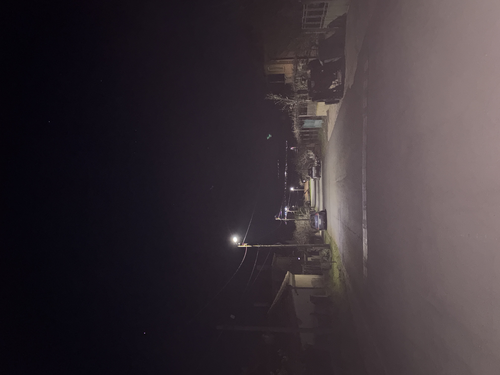Night scene in Goritsa, Bulgaria. A street with a single car parked on the right side. Streetlights illuminate the road. Houses are visible along the street. The sky is dark.