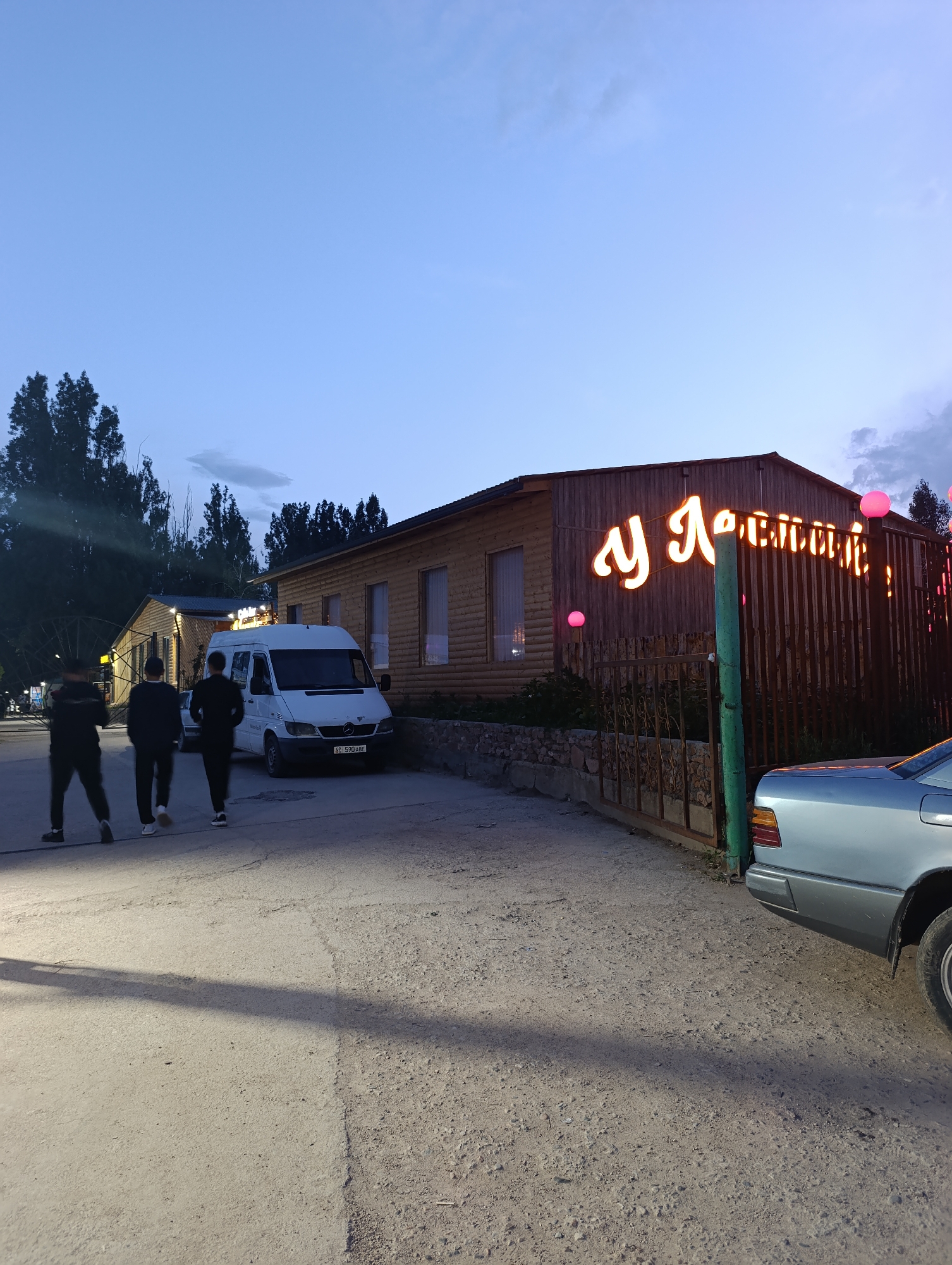 The image depicts an exterior scene in Bosteri, Kyrgyzstan, at dusk. In the foreground, three individuals, two walking away from the camera and one facing partially away, are on a gravel pathway. Behind them, a white Mercedes-Benz Sprinter van is parked. To the right, a light blue Mercedes-Benz E-Class sedan is partially visible. The main subject is a wooden building with a fence in front. A prominent neon sign with Cyrillic lettering glows on the building's facade. The sky above is a soft blue with hints of clouds. The overall ambiance suggests a relatively quiet evening in a semi-urban or recreational area.
