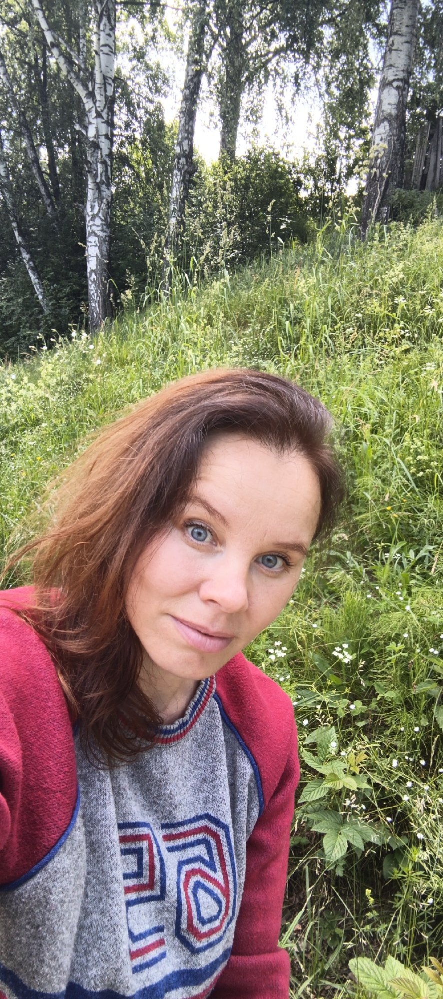 A woman with reddish-brown hair and blue eyes is pictured in a selfie, seated outdoors in a grassy, wooded area. She is wearing a gray sweater with a red and blue geometric design and a red cardigan. The background features birch trees and lush green grass with small white wildflowers. The location is the city of Chaikovsky, Russia.