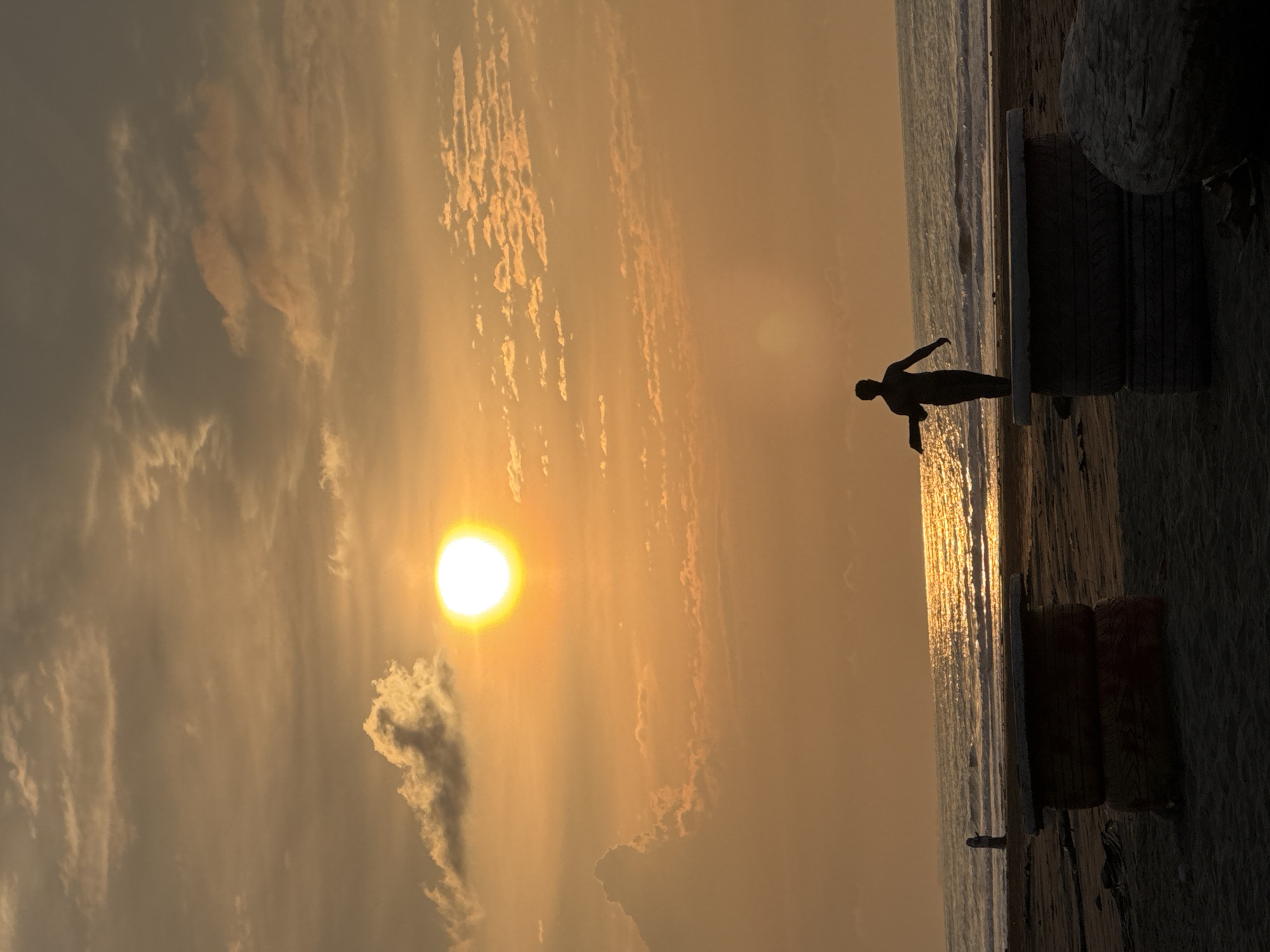 A silhouette of a person stands on a makeshift bench on a beach, facing the ocean at sunset. The sky is filled with an orange glow and scattered clouds. The ocean reflects the sunlight, creating a shimmering path. In the foreground, two stacked tires form the base of the bench. A person is visible in the distance, standing near the water. The scene appears to be on a beach in Reudeuep, Indonesia.