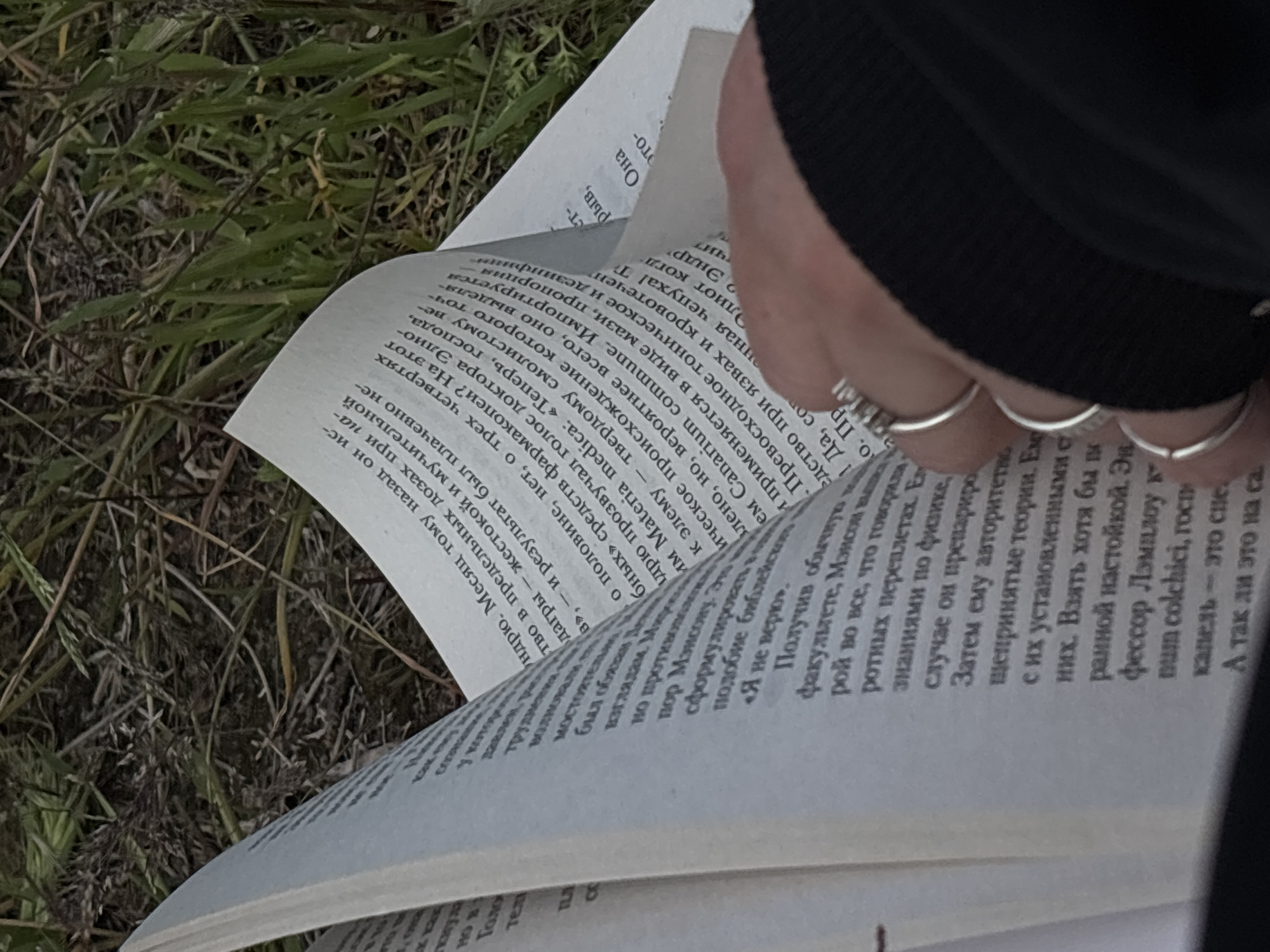 Here is a detailed description of the image:
**Scene:** An open book rests on the ground, amidst grass and other low-lying vegetation. A hand with silver rings is turning the page. The text on the open pages is in Russian. The environment appears to be outdoors.