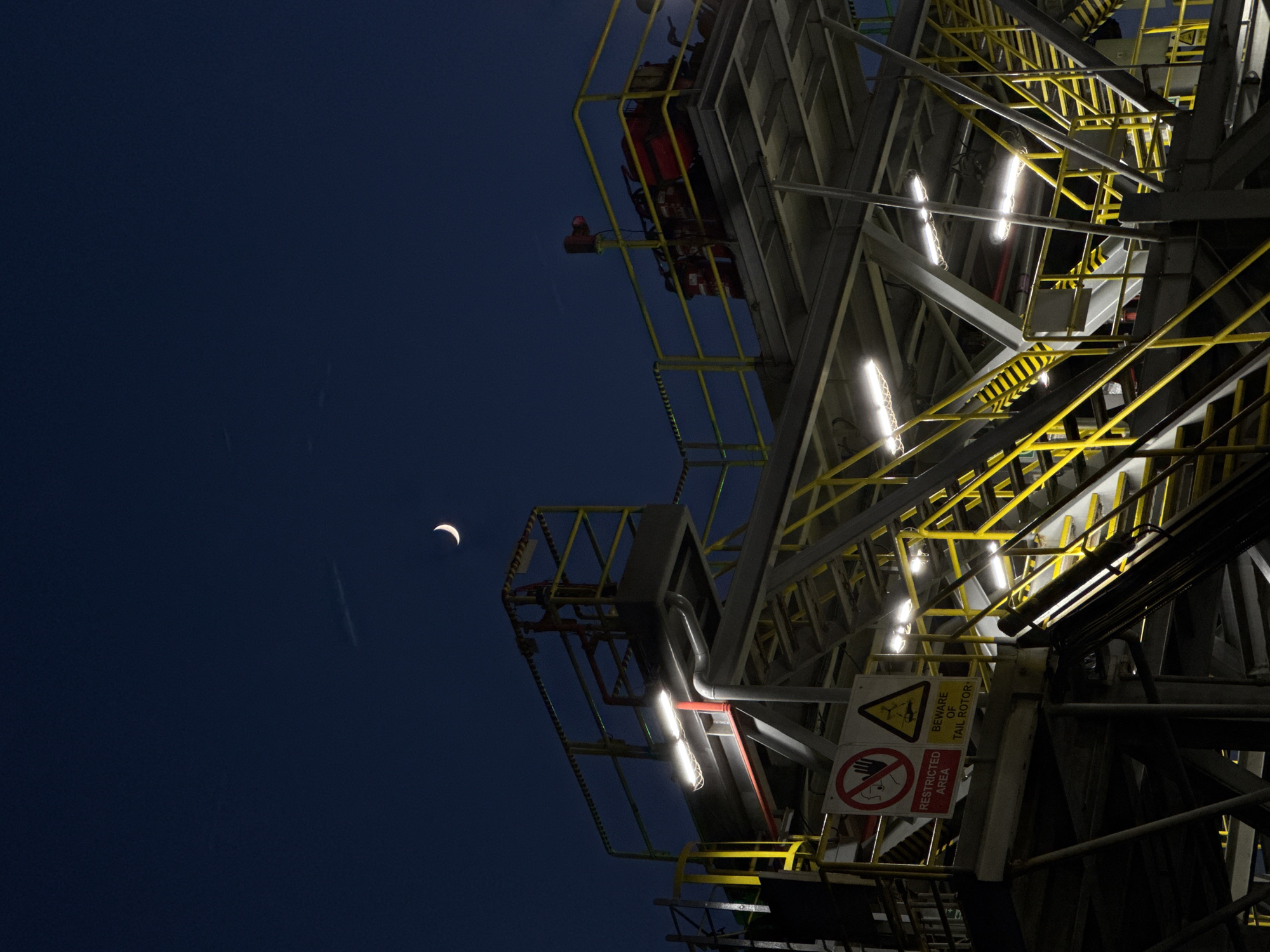 The image depicts an industrial structure at night, possibly an oil rig or a similar facility, with a crescent moon visible in the deep blue, twilight sky. The structure features multiple levels with bright yellow railings and staircases. Several lights illuminate the walkways and stairs. Two prominent signs are visible: one red and circular with a white bar and a figure with a hand, indicating "RESTRICTED AREA," and another triangular yellow sign with a black symbol and text that reads "BEWARE OF TAIL ROTOR." Red fire extinguishers are also visible on an upper platform. The overall scene is one of industrial activity under a darkening sky. The location is described as Unknown, Italy, but no specific urban features are visible.