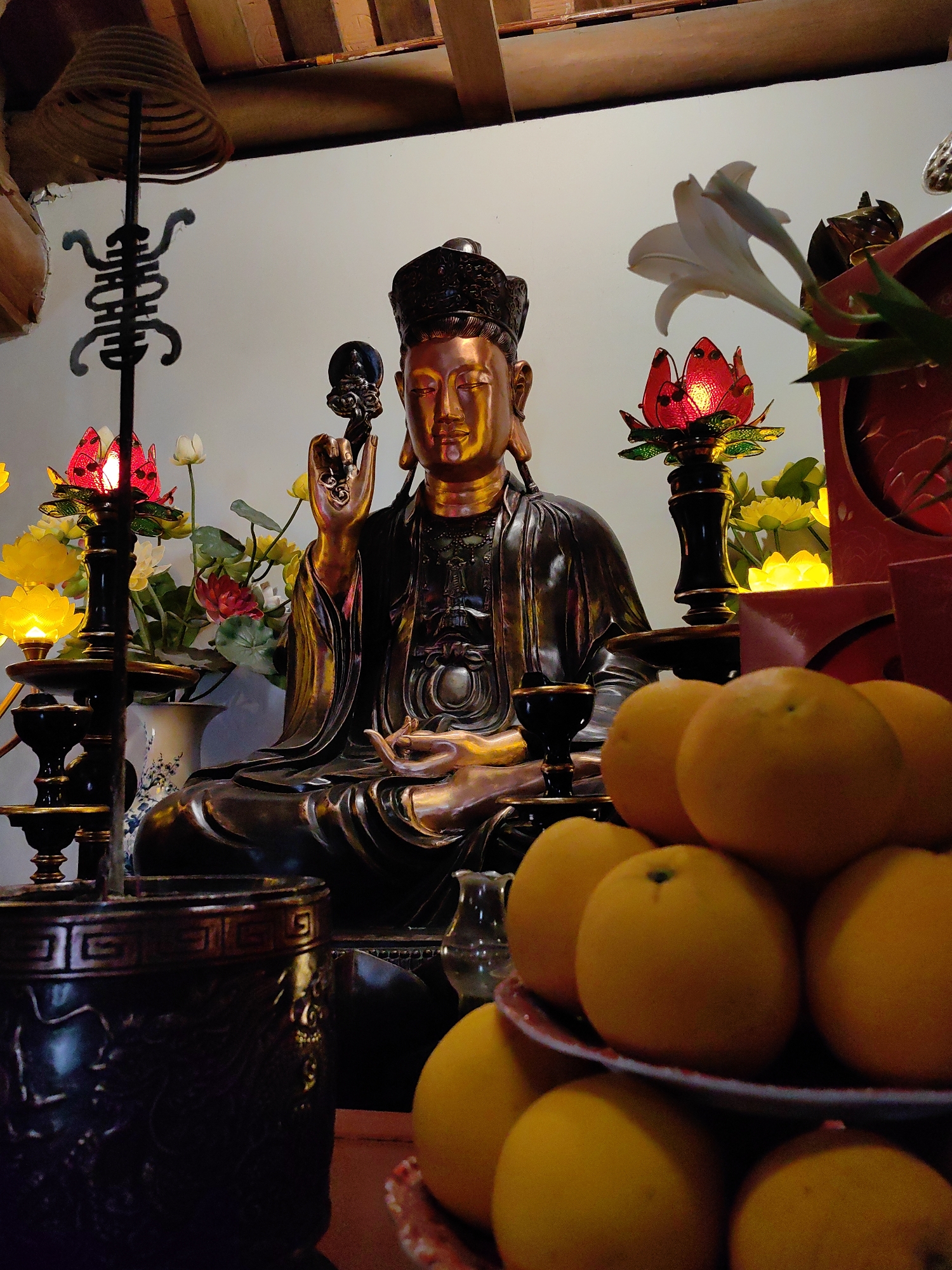 The image depicts an altar in what appears to be a religious or domestic setting, possibly in Hong Gai Ward, Vietnam. In the center is a bronze-colored statue of a seated figure wearing an elaborate headdress, likely a representation of a deity or religious figure. The figure holds an object in its raised right hand. The altar is adorned with various items, including flowers (both real and artificial), red lotus-shaped candle holders, and a plate of oranges in the foreground. To the left of the statue is an incense coil hanging from a stand, and to the right is a small red shrine. The background is a plain white wall. The lighting is soft and ambient, creating a reverent atmosphere.