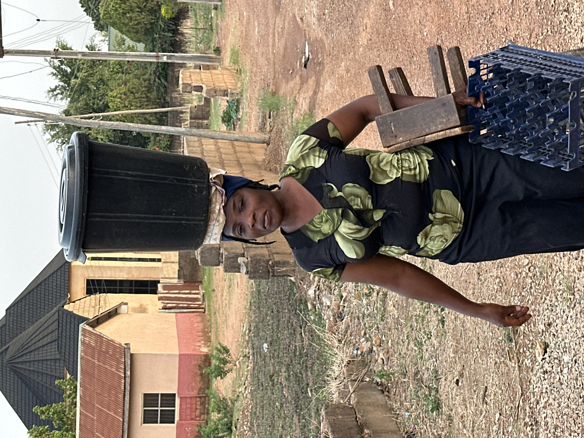 A woman stands outdoors, skillfully balancing a large, black plastic basin on her head. She wears a black top with a green floral or leaf pattern and dark trousers. A blue headwrap is visible beneath the basin, supporting the load. In her left hand, she holds a small, wooden stool, while her right hand carries an inverted blue plastic crate, possibly for bottles or other goods. Her facial expression is neutral as she looks towards the camera.
The scene unfolds in what appears to be a rural or semi-urban setting. The ground is a mix of reddish-brown dirt and gravel, with sparse, dry vegetation and scattered rocks. In the background, several simple buildings are visible, some with corrugated iron roofs and others with dark tiled roofs. Utility poles with power lines stretch across the sky, and green trees and dense foliage are seen in the distance. The lighting suggests it is daytime. The image captures an everyday activity of transporting items, typical of life in communities like Bukuru, Nigeria. There is no visible text in the image.