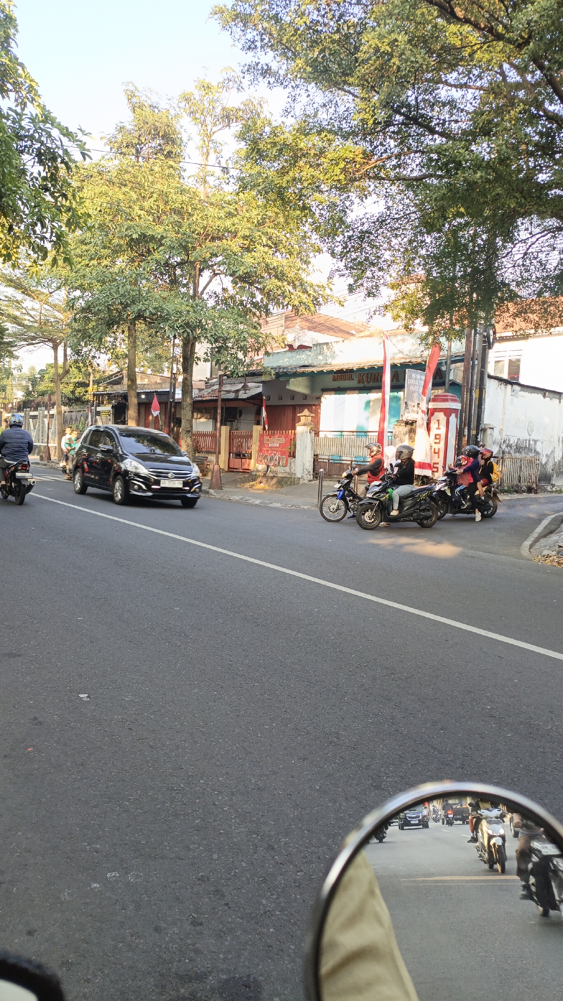 The image captures a street scene in Salatiga, Indonesia, with a focus on traffic and local businesses. The foreground shows the perspective from a vehicle, with a side mirror reflecting traffic and a portion of the driver's leg. The road is paved with asphalt, and a white line marks the lane. On the right side of the road, a building with a white facade and a patterned roof is visible, along with a red and white striped pole that has "1945" inscribed on it. Red and white flags, typical of Indonesian Independence Day, are displayed. Several motorcycles are present, some with riders wearing helmets. One motorcycle is seen in the foreground reflection, and other motorcycles are on the road. A black car is moving in the left lane. Trees with green foliage line the left side of the street, and the sky is a clear light blue. Buildings with shops are visible behind the trees, some with signs that are partially obscured. The overall impression is a busy urban street during daylight hours, likely in the morning or late afternoon given the lighting.