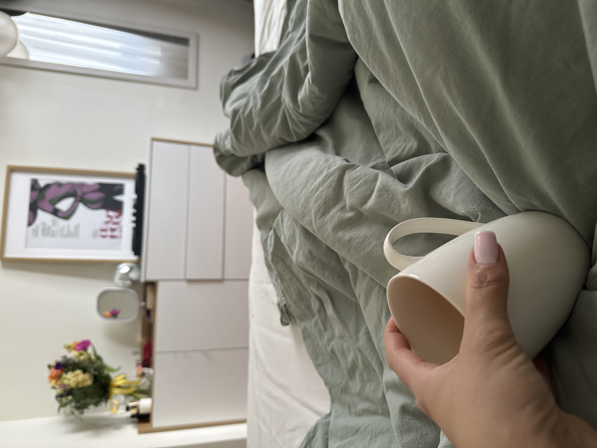 A first-person view in an indoor setting. A hand holds a white mug with a handle. The backdrop includes a bed covered with an olive green duvet, against a wall with white cabinetry. A framed art print hangs above a vanity with a mirror and a bouquet of flowers. The location is not explicitly defined in the image. The location in the city of Belgrade, Serbia cannot be determined.