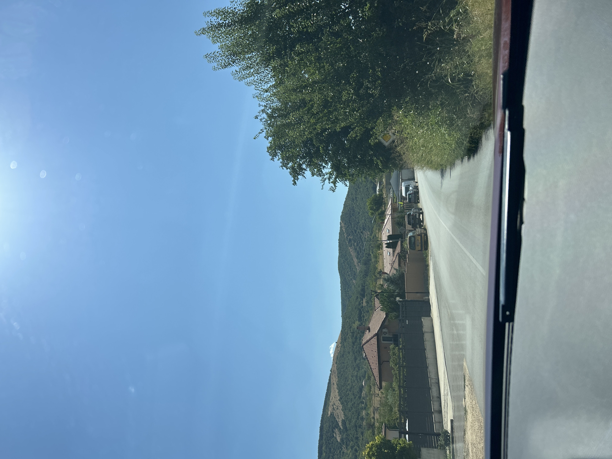This is a roadside view from inside a vehicle looking out at a clear, sunny day in Markovo, Bulgaria. The road, paved with asphalt and marked with a white line, curves slightly to the right. On the left side of the road, there are several houses with red tiled roofs, fences, and vegetation. The houses appear to be residential properties. Further in the distance, rolling hills covered in trees and dry grass form the backdrop.
On the right side of the road, a large, leafy green tree dominates the foreground, casting shadows on the road. Beyond the tree, there is a patch of tall grass and wildflowers. A yellow diamond-shaped road sign with a black symbol is visible on a post near the tree.
In the middle of the road, several vehicles are present. There is a white van, a dark-colored pickup truck, and another light-colored vehicle. The sky is a bright, clear blue with a few faint white clouds and lens flares from the sun, which is high and bright. The perspective suggests the image was taken from the driver's or front passenger's seat of a car, with the windshield wipers and part of the car's dashboard visible in the foreground. The overall scene depicts a rural or semi-rural environment on a summer day.