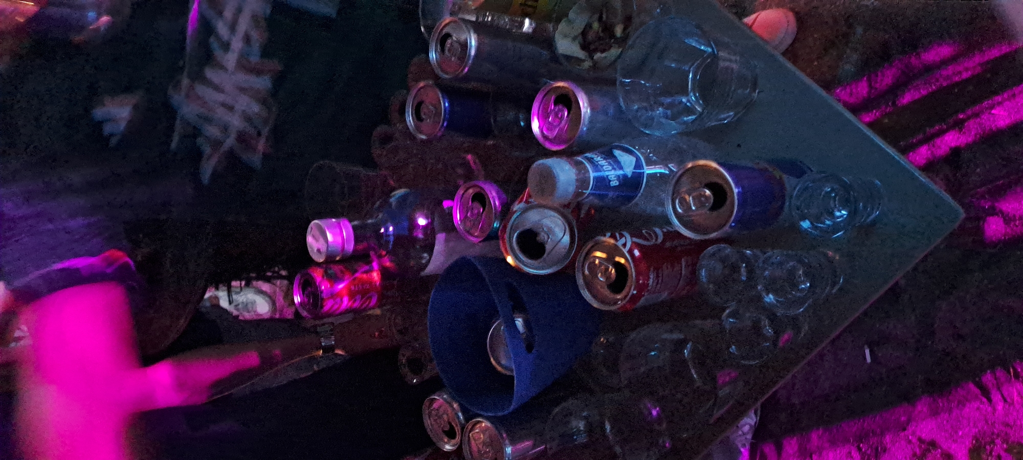 The image depicts a table littered with empty drink cans and bottles, suggesting a party or gathering. The scene is illuminated by pink and purple lights, creating a vibrant, possibly nighttime atmosphere. Several empty beer cans, some red and some blue, are scattered across the table. A blue bucket, likely for ice or trash, is also present, containing a few cans. A bottle with a silver cap and a blue label is visible. The table itself is dark and appears to be made of a reflective material. In the background, the scene is dark with blurry elements, possibly indicating other people or objects. The overall impression is one of a messy but lively post-event setting. The requested information about Burgas, Bulgaria cannot be determined from the image.