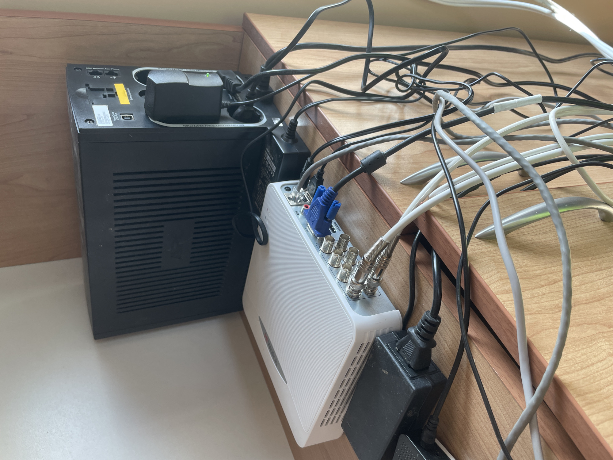 This media file captures an indoor, close-up view of electronic equipment and tangled cables, likely part of a surveillance system setup. The scene is set on a light wooden surface with a darker wooden trim in the background, suggesting a desk, shelf, or cabinet.
The primary subjects are a black Uninterruptible Power Supply (UPS) unit, a white Digital Video Recorder (DVR) or Network Video Recorder (NVR), and numerous power adapters and cables. The black UPS unit, positioned on the left, features several ports on its top surface, including a USB port and connections labeled "Dsl / Modem / Phone" for surge protection. Text "Battery Backup + Surge Protection" and "SURGE PROTECTION" are visible. A yellow "CAUTION" sticker is also partly seen. A power adapter with a green LED indicating power is plugged into the UPS.
To the right of the UPS, the white DVR/NVR unit is visible. Its rear panel shows multiple coaxial (BNC) input connectors, a blue VGA output port, and a partially obscured HDMI port. Several coaxial cables are connected to these inputs. A power adapter, possibly branded "CEON," powers this device.
A significant detail is the extensive tangle of cables covering the wooden surface. These include black power cables, grey and white data or signal cables (possibly Ethernet or additional video lines), and the blue VGA cable connecting to the DVR. The overall impression is one of disarray due to poor cable management. The lighting suggests it is daytime. The equipment appears functional and interconnected, typical of a security camera system installation.