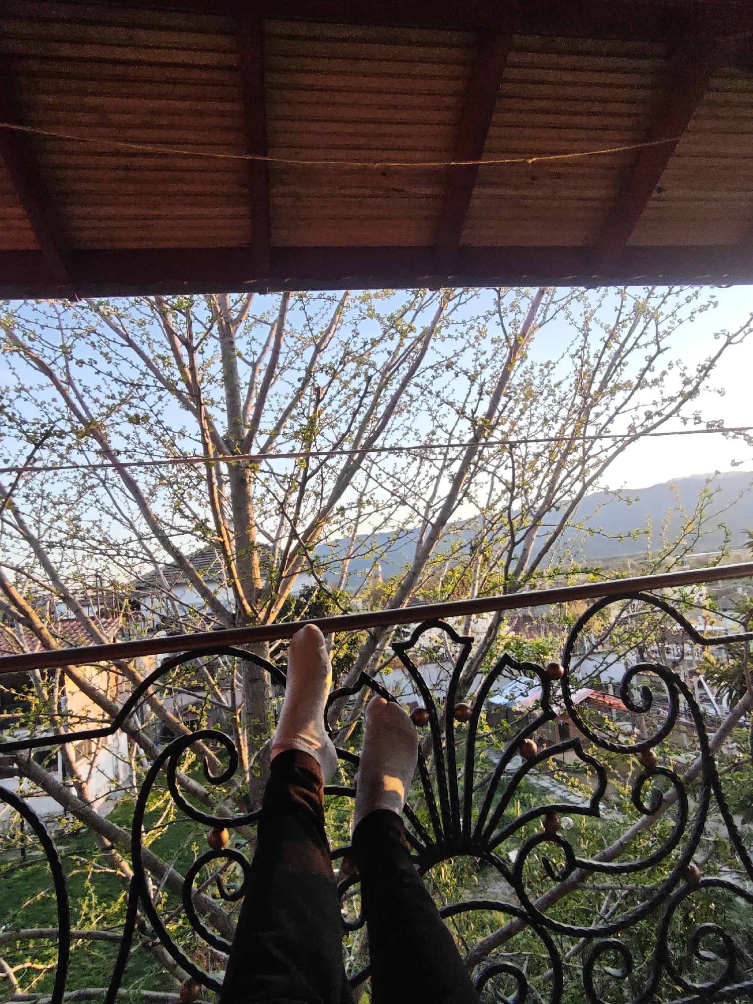A first-person perspective photograph shows a person's legs and feet, clad in dark trousers and white socks, propped up on a black, ornate wrought iron balcony railing. Beyond the railing is a view of tree branches, some with budding leaves, against a partly cloudy blue sky. Houses and hints of rooftops can be seen through the branches, suggesting an urban or suburban setting. A distant mountain range is visible on the right side of the image. Above the scene, part of a wooden ceiling or overhang is visible, which could be part of the balcony structure.