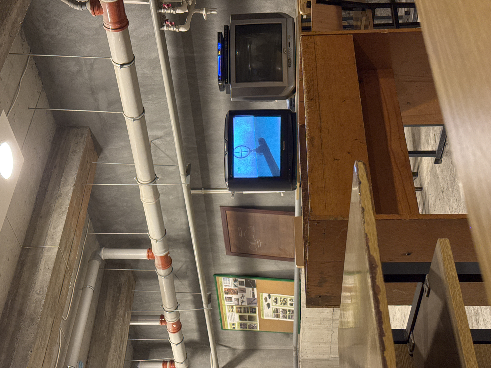 The image shows an indoor scene with a focus on pipes running across a concrete ceiling. Attached to the ceiling and to a wall, there are two cathode-ray tube (CRT) televisions. Below the televisions, a bulletin board and a framed piece of paper are affixed to the wall. A wooden cabinet or shelving unit is prominent on the right. The pipes are supported by metal cables. The image suggests an older, possibly industrial or institutional setting. Based on the prompt, the location is likely Yerevan, Armenia.