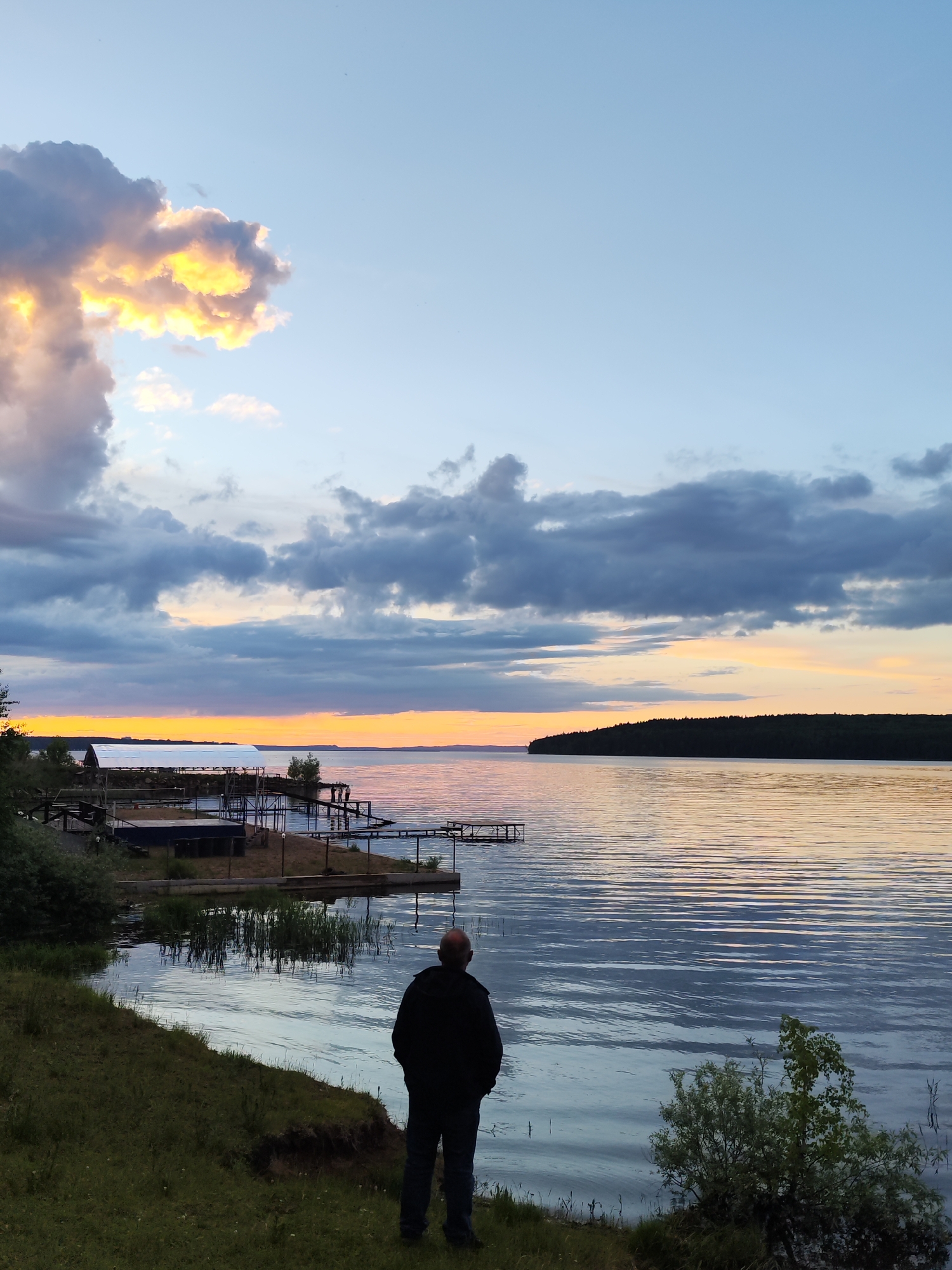 The image shows a man standing on a grassy bank, looking out over a body of water. The water appears calm, with horizontal ripples. In the distance, there is a dark, tree-covered landmass on the other side of the water. To the left, there are structures that appear to be docks or boathouses along the shoreline. The sky is filled with clouds, with the setting sun casting a warm, golden light on some of the clouds. The man is wearing dark clothing and is facing away from the camera.
The location is the city of Chaikovsky, Russia.