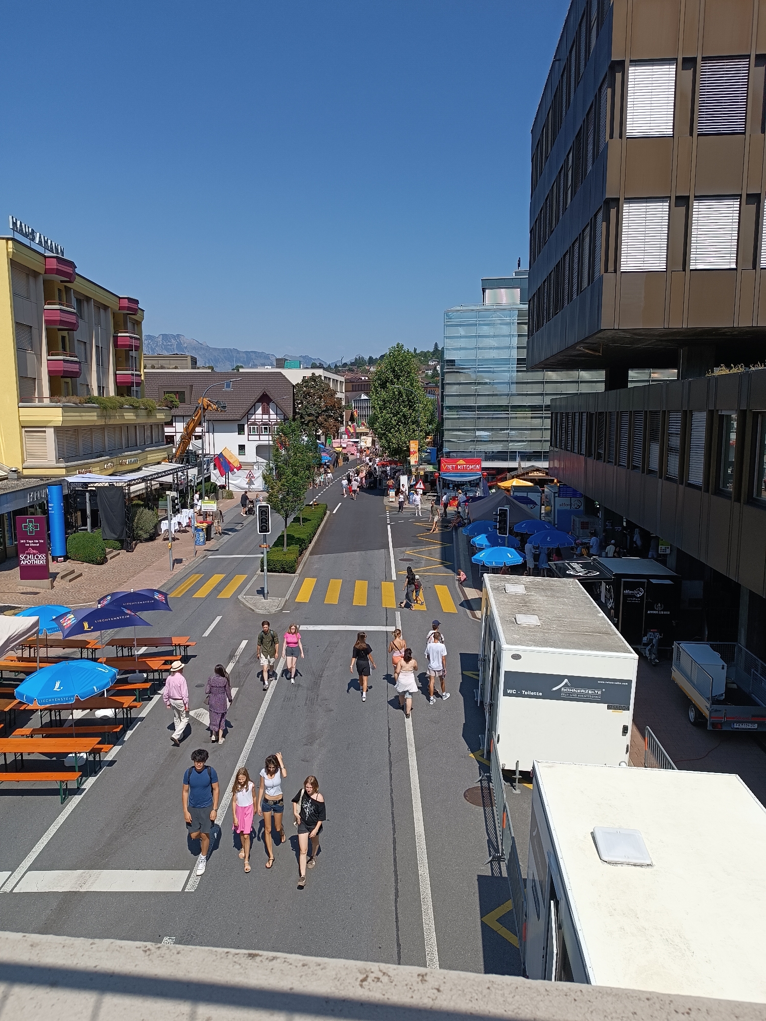 The image depicts a street scene in Vaduz, Liechtenstein, on a clear, sunny day. The perspective is from a slightly elevated viewpoint, looking down onto a street lined with buildings and an outdoor gathering or event.

In the foreground, there are several picnic tables with blue umbrellas, suggesting an outdoor seating area for an event. A white portable toilet unit with "RÖHNER ZELTE" branding is visible on the right side of the road.

The street is bustling with pedestrians. People are seen walking in various directions, some in small groups, others individually. They are dressed casually, indicating warm weather. Clothing items visible include t-shirts, shorts, dresses, and hats.

The environment includes commercial buildings with storefronts and signage, a glass-fronted modern building, and a multi-story building with a distinctive brown facade and rows of windows. Trees are planted along the side of the street. In the background, mountains are visible under a clear blue sky.

A traffic light is present at an intersection. Yellow crosswalk markings are clearly visible on the asphalt. Several tents and temporary structures are set up along the sidewalks, suggesting a market, festival, or other public event. A crane is visible on one of the buildings, possibly indicating ongoing construction or event setup.

The overall scene portrays a vibrant street life with people enjoying an outdoor event in a European town setting.