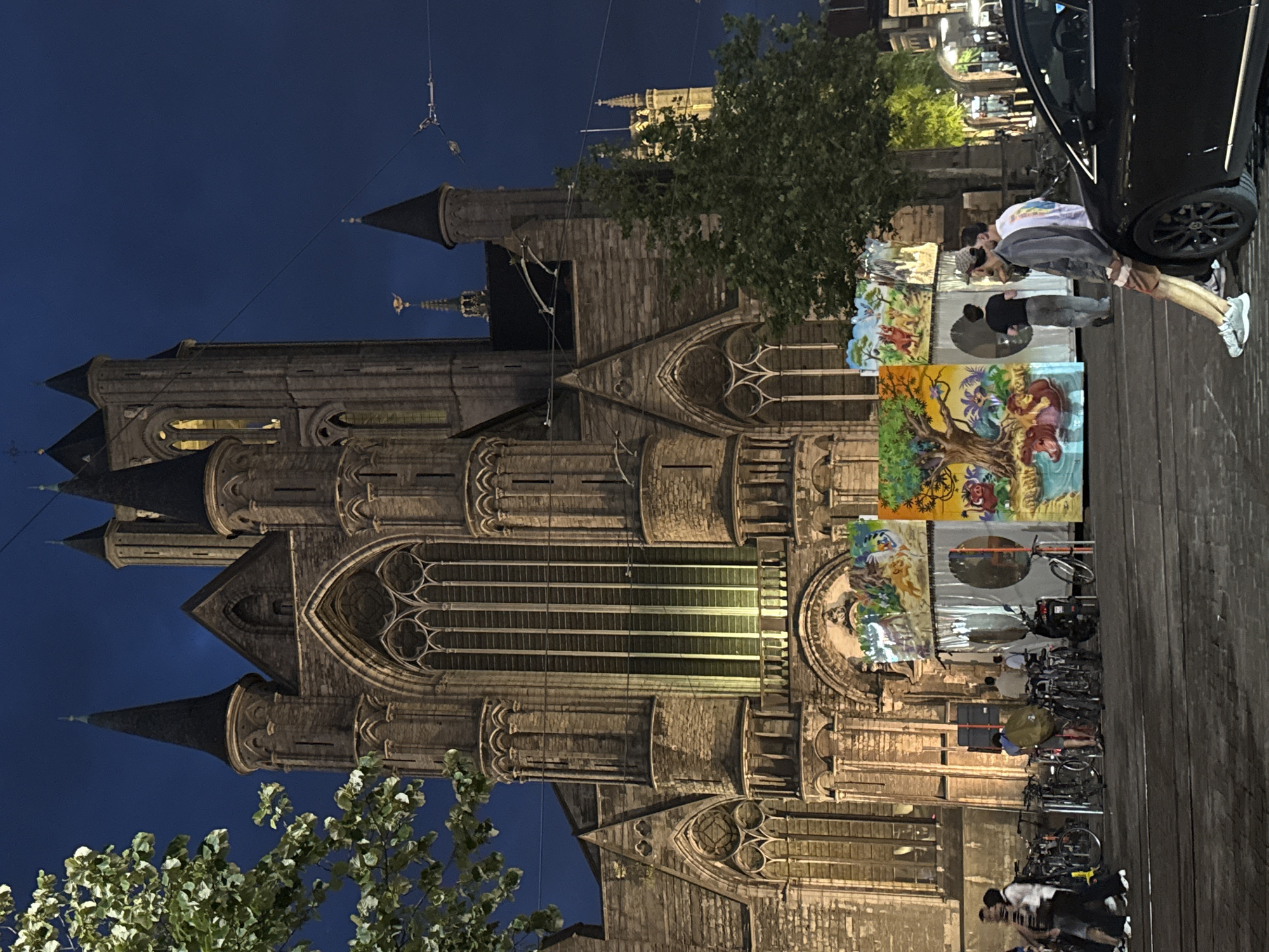 The image captures a nighttime scene in the city of Ghent, Belgium, with the imposing Saint Nicholas' Church (Sint-Niklaaskerk) as the dominant backdrop. The church, a magnificent example of Gothic architecture, is illuminated, highlighting its intricate stonework, pointed spires, and large stained-glass windows. In the foreground, several people are visible. Some are standing near a row of parked bicycles and a scooter, while others are gathered around a colorful temporary structure adorned with cartoonish depictions of animals, reminiscent of a jungle or safari theme. The ground appears to be paved, with streetlights and overhead wires suggesting an urban environment. A black car is parked on the right side of the frame, partially obscuring the view of the street. The overall atmosphere is that of a lively public space at dusk or night.