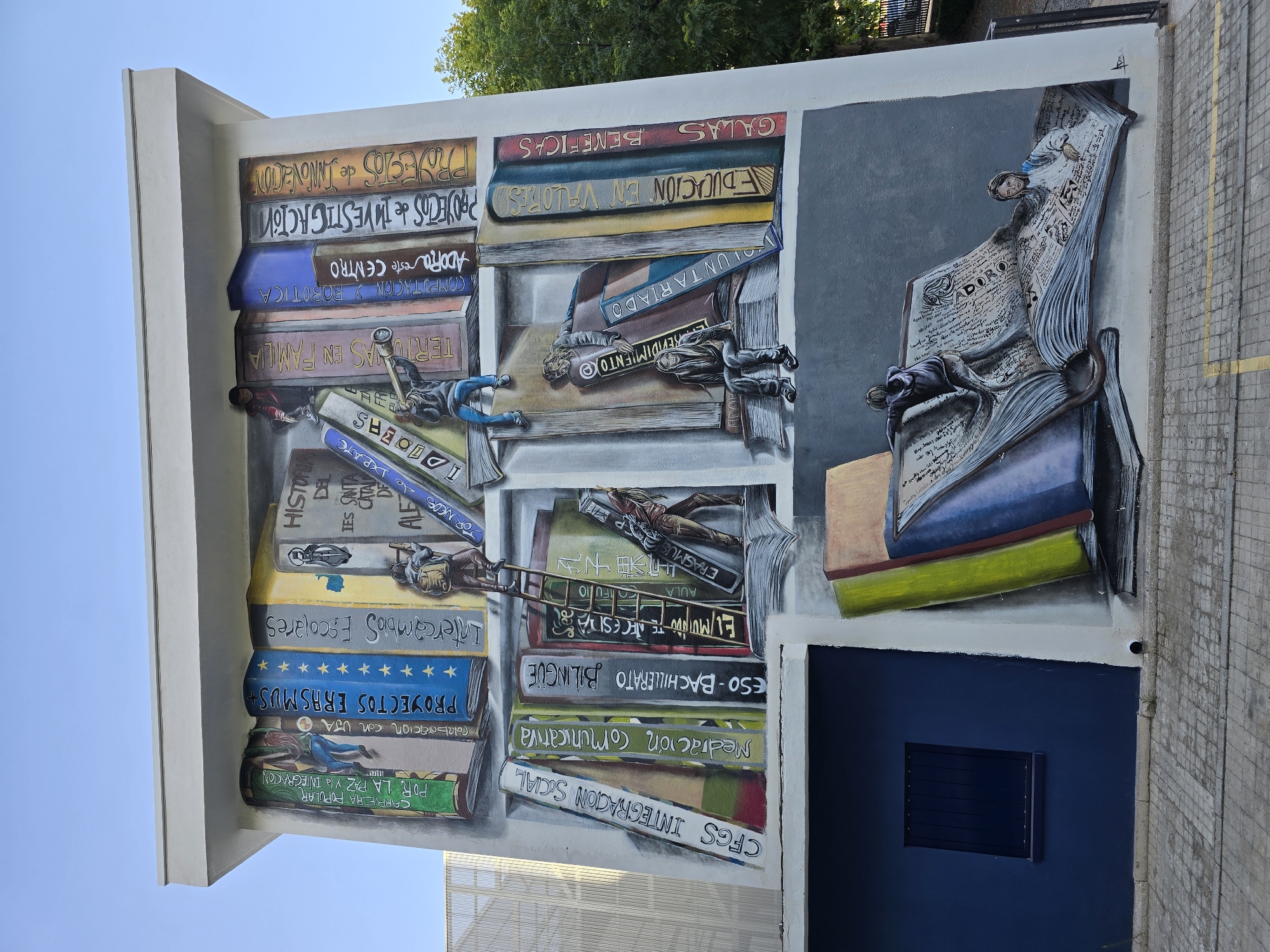 This is a mural on the side of a building, depicting a large bookshelf filled with books and people interacting with them. The mural is painted to look like a wall of books, with titles such as "CARRERA POPULAR POR LA PAZ Y LA INTEGRACION," "PROYECTOS ERASMUS," "Intercambios Escolares," "HISTORIA DEL IES SANTA CATALINA DE ALEJANDRIA," "CFGS INTEGRACION SOCIAL," "Mediacion Comunicativa," "ESO - BACHILLERATO BILINGUE," "EL MUNDO TE NECESITA," "AULA ERASMUS," "Voluntariado," "EDUCACION EN VALORES," and "GALAS BENEFICAS."
Several figures are depicted climbing on, reaching for, and reading the books. One person is using a telescope to look at a book, while another is climbing a ladder. Two people are shown on a giant open book at the bottom of the mural, one seemingly reading and the other climbing out. The building itself is white with a light grey trim, and a dark blue section with a window is visible on the left. The ground in front of the mural is paved with interlocking bricks. Trees are visible in the background on the right side of the image. The sky is clear and blue.