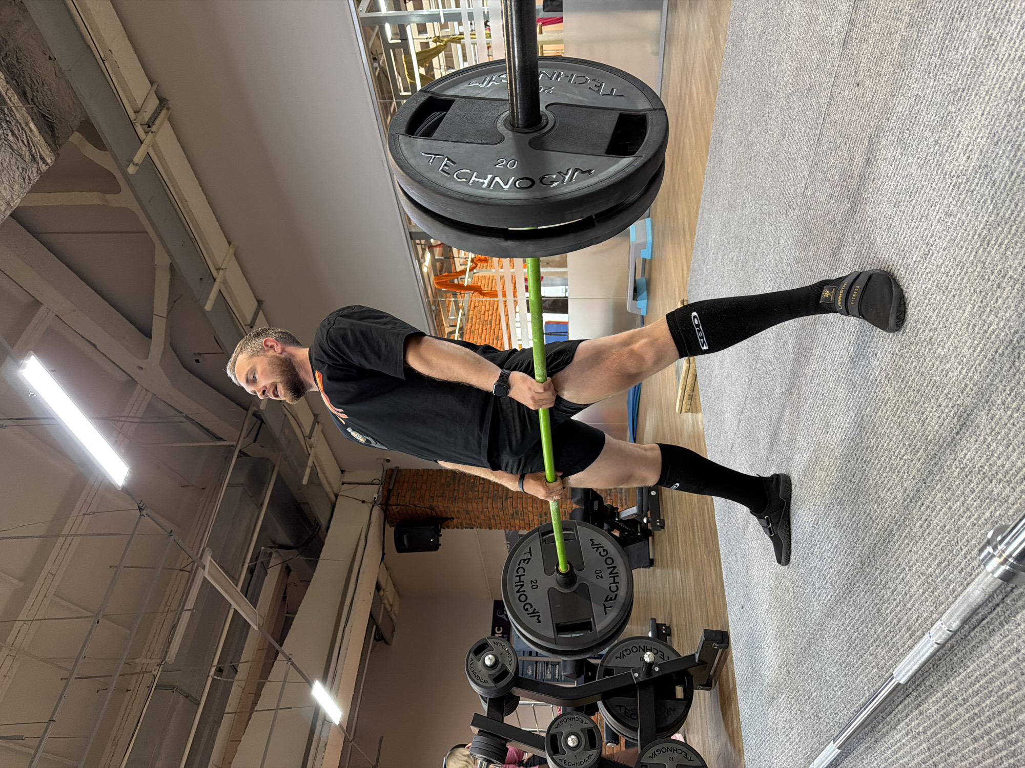 A man in a black t-shirt and black shorts, knee-high socks, and black athletic shoes is standing in a gym, holding a loaded barbell with a green stick-like attachment that extends from the barbell. The barbell has two large black weight plates labeled "TECHNOGYM 20" on each side. The man has a beard and is looking to his left. The gym has grey carpet and weights and weight storage equipment are visible. The gym is in Chaikovsky, Russia.