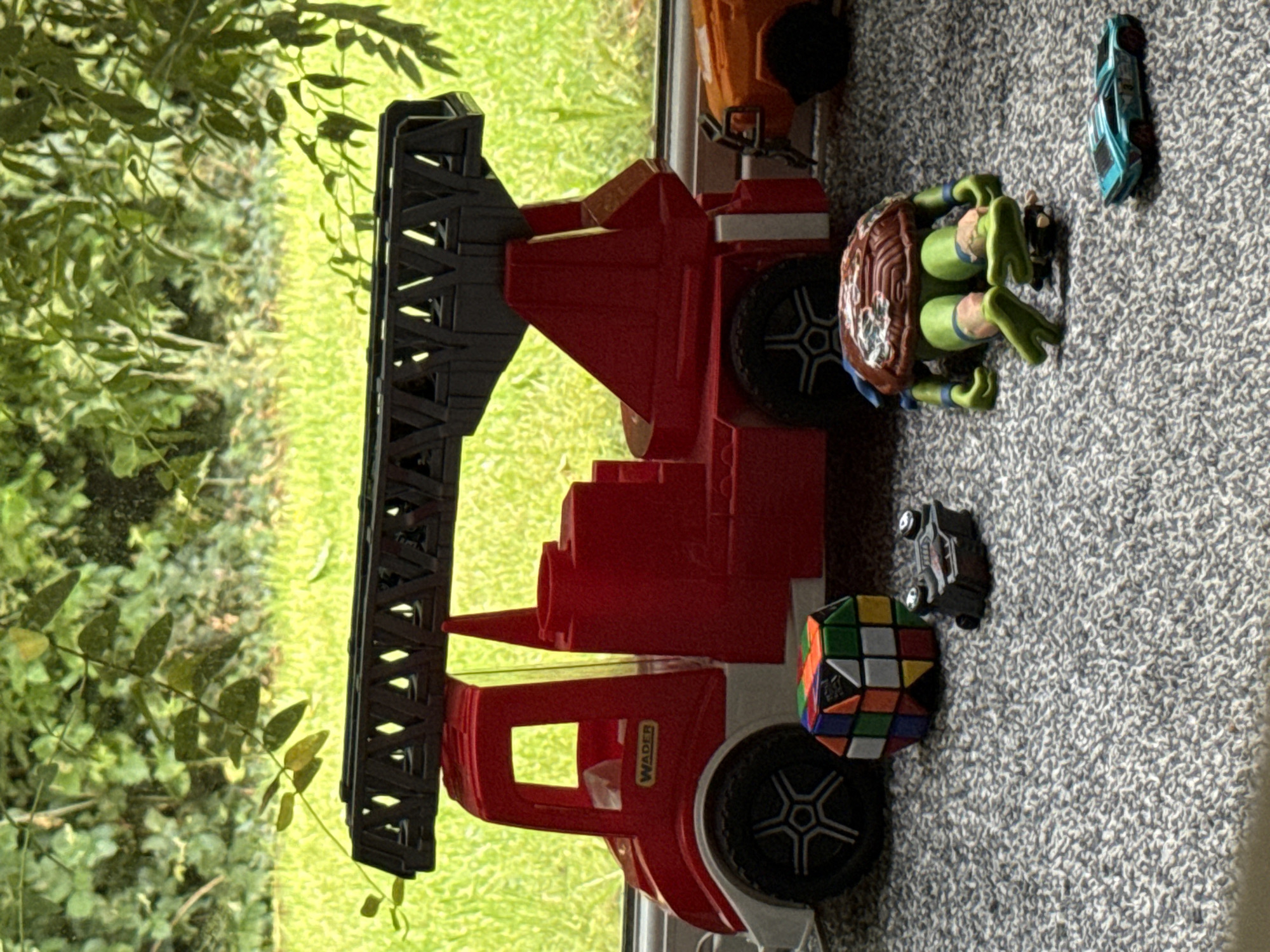 The image captures a collection of children's toys arranged on a grey, textured carpet next to a window, looking out onto a vibrant green outdoor scene, likely in Hazlemere, United Kingdom.
The main subject is a large, red toy fire truck with a black extendable ladder. The truck features white or light grey accents and black wheels. The brand name "WADER" is clearly visible in yellow text on the side of the truck's cab, above the front wheel.
To the right of the fire truck, a green Teenage Mutant Ninja Turtle action figure with a brown shell is positioned on the carpet, appearing to be in a crouching or resting pose. Nearby, a partially scrambled Rubik's Cube, displaying various colors including red, green, blue, yellow, white, orange, and black, rests near one of the fire truck's front wheels. A small black toy car is also visible near the Rubik's Cube. Further to the right, a larger blue and green toy car is situated on the carpet, and part of an orange or brown toy vehicle is visible near the top edge of the frame, behind the fire truck.
The outdoor background, seen through the window, is filled with lush green foliage, suggesting trees or bushes, and a bright green grassy area, indicating a clear, bright day. The light from outside illuminates the scene. There is no explicit activity taking place; the toys appear to be set up or simply resting on the carpet.