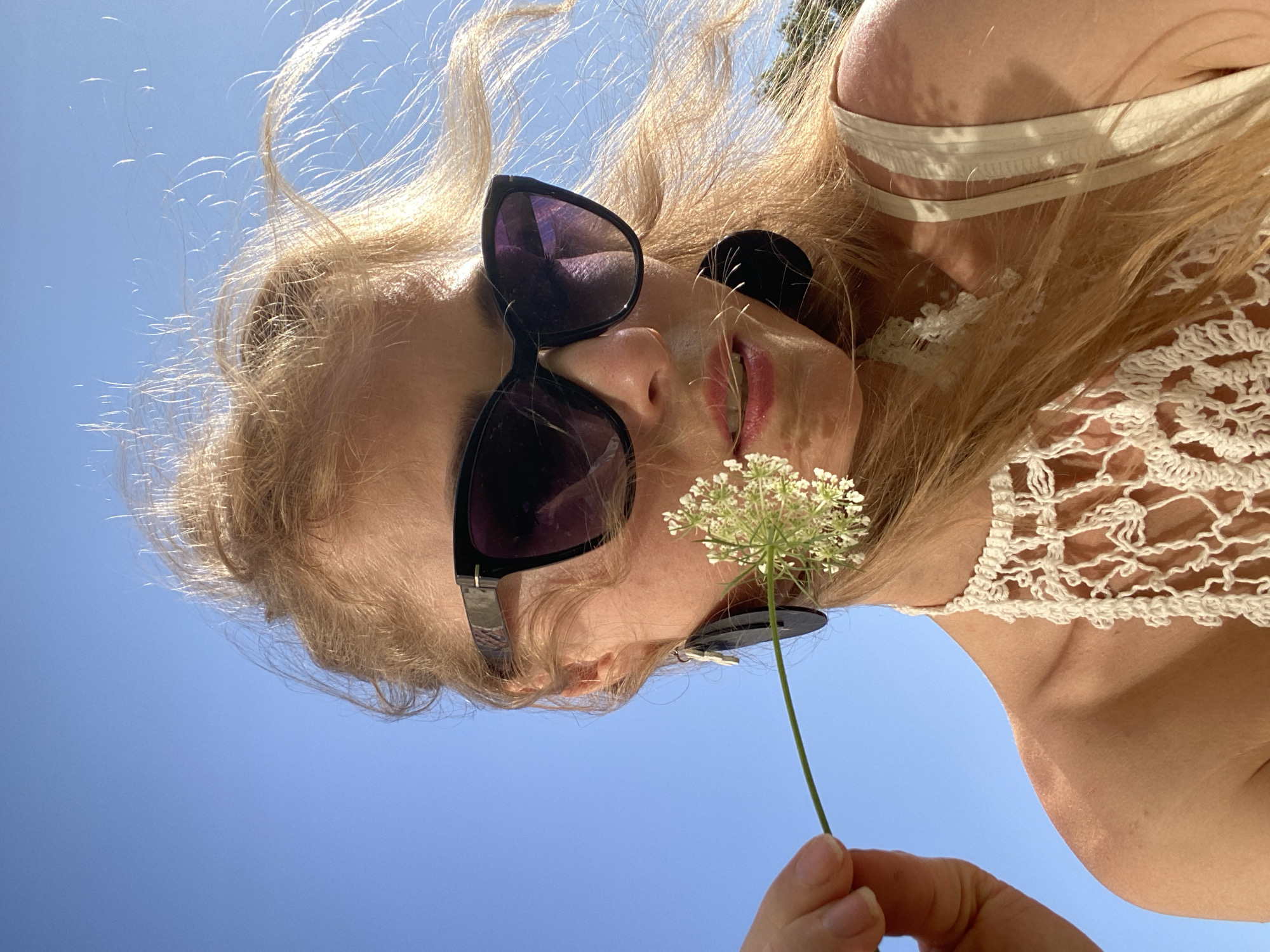 A close-up, low-angle shot captures a blonde woman in sunglasses holding a white wildflower to her lips. The woman, with her hair blowing in the wind, wears a cream-colored crochet top and large, dark earrings. The background is a clear blue sky, suggesting an outdoor setting, possibly in the city of Lida, Belarus. The lighting indicates it is daytime, with the sun casting a warm glow on the subject.