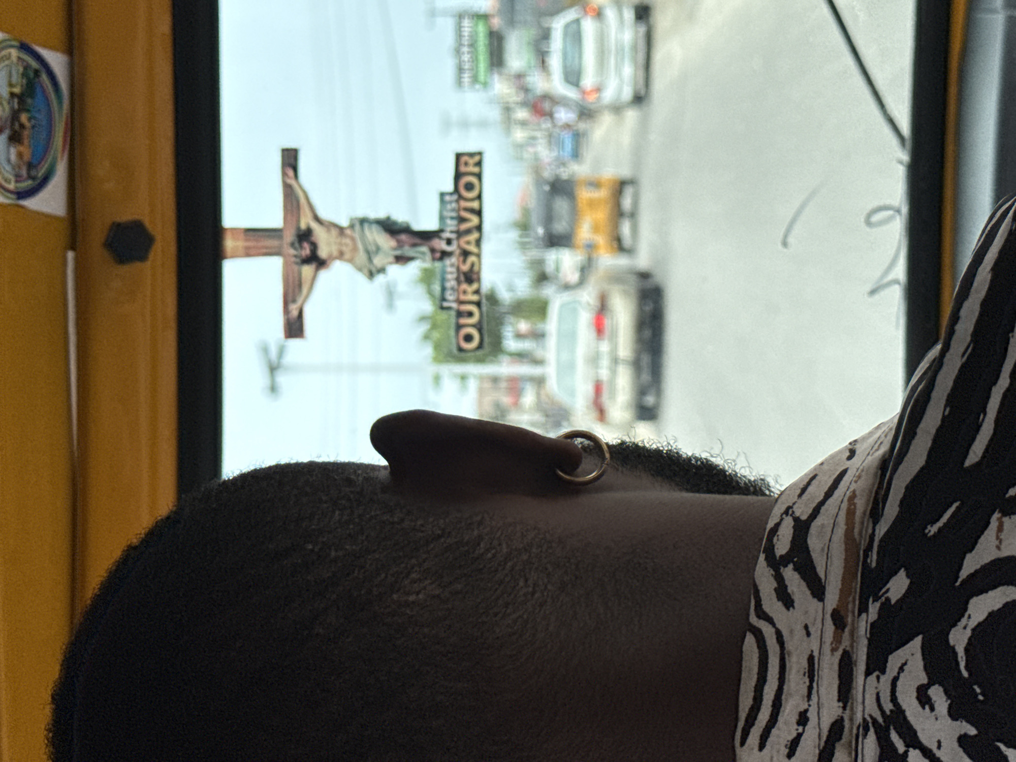 The image shows a close-up of a person's head with dark, short hair and a gold hoop earring in their ear. The person is wearing a black and white patterned shirt. A window is visible in the background, showing a street scene in Ado, Nigeria. Through the window, there's a crucifix with Jesus, a sign that reads "Jesus Christ Our Savior," cars, and other street elements. A sticker is attached to the window.