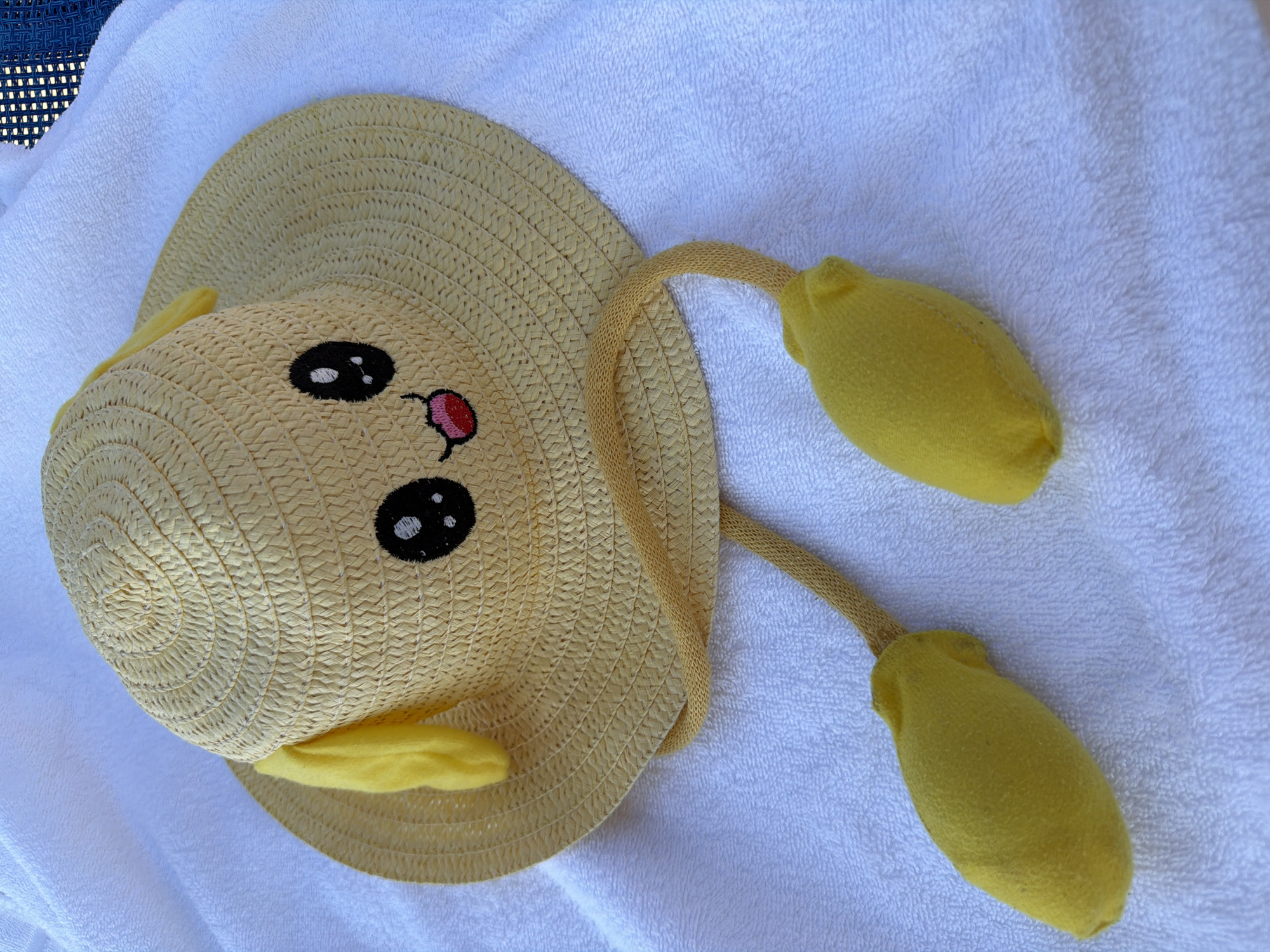 A straw hat with a cartoonish face and dangling yellow ear-like appendages is laid on a white towel. The hat is yellow and woven, with black eyes, a pink tongue, and two yellow felt ears protruding from the sides. Two yellow cord-like strings, each ending in a plush yellow stuffed element, hang from the hat and rest on the towel. The towel itself is textured and appears to be lying on an outdoor surface, possibly near a pool or beach area, given the presence of a dark blue chair with a woven pattern visible in the upper right corner. The scene suggests a relaxed, summery atmosphere. The location is identified as Gatteo, Italy.
