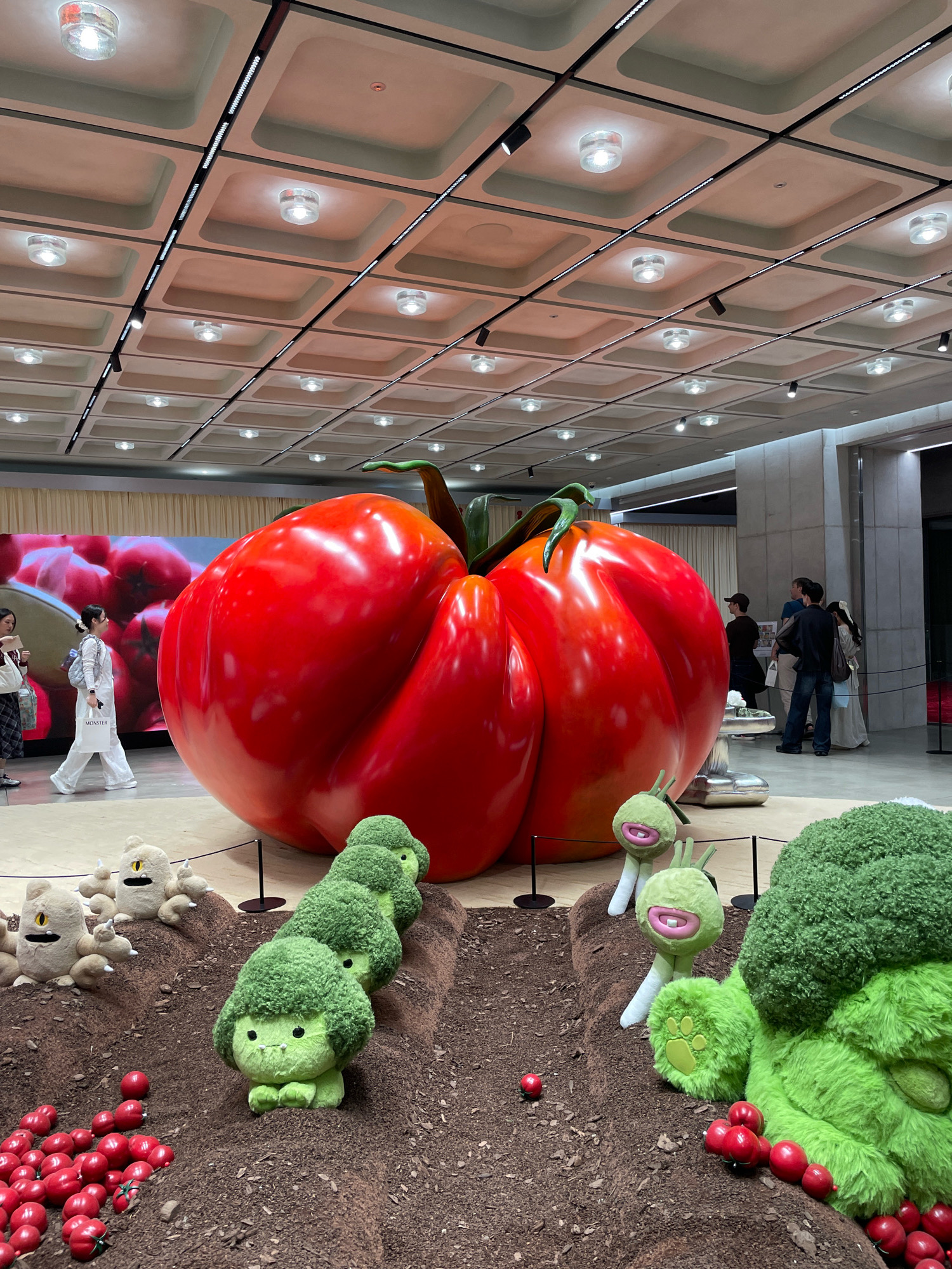 The image captures a whimsical indoor display that appears to be at a public venue or exhibition space in Seoul, South Korea. Dominating the scene is a colossal, bright red tomato sculpture, gleaming under artificial lights. In the foreground, a miniature landscape is set up with dark brown soil, featuring a row of adorable plush broccoli characters with green, fluffy heads and cute faces. To the left, several tan, fluffy plush creatures with single yellow eyes and black mouths are scattered in the "soil." Red, berry-like objects are also distributed across the ground, mimicking tomatoes or similar fruits.
In the background, a large screen displays an image of more red fruits, likely contributing to the overall theme. Several individuals are present, observing the display. On the left, two women are walking, one in white attire carrying a shopping bag. On the right, a group of people are gathered, some looking towards the main sculpture. The ceiling features a grid of square panels with recessed lighting. The overall ambiance is playful and visually striking, suggesting an art installation or a themed promotional event.