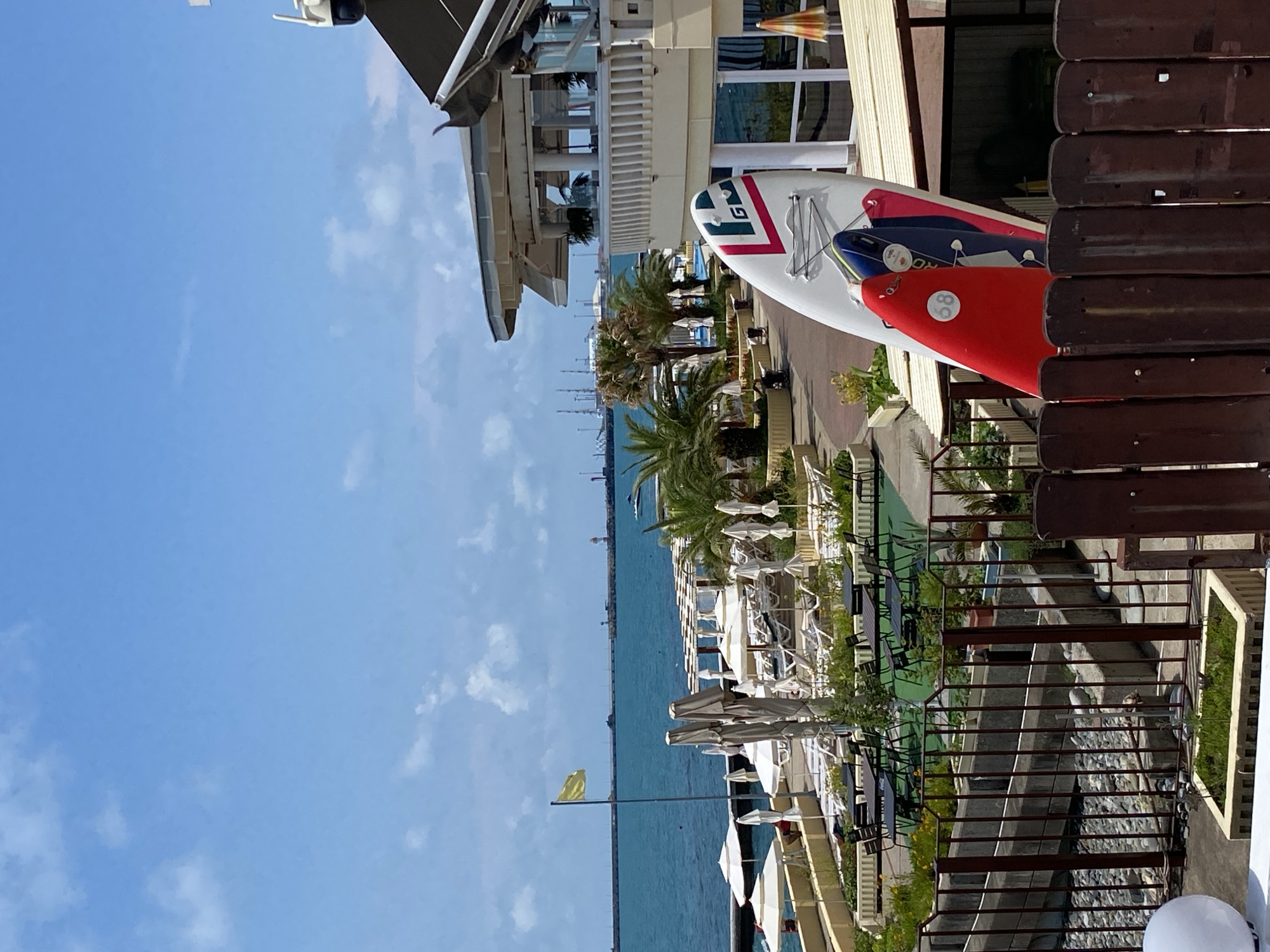 This media file captures a vibrant scene likely in Sochi, Russia, on a bright, sunny day. The image is oriented vertically, showcasing a waterfront promenade with elements of a resort or leisure area.
Dominating the foreground and midground are several surfboards, one prominently red with a white circular marking containing the number "88", and another a larger white board with colorful stripes and a "GM" logo. These suggest recreational activities related to water sports.
Beyond the surfboards, a row of white umbrellas and lounge chairs hints at a beach or pool area. Palm trees interspersed with the seating further reinforce a warm-climate, resort setting. The calm, blue sea stretches out to the horizon, where a long pier or breakwater is visible, along with a faint lighthouse or structure in the distance.
The architecture of the buildings suggests a modern establishment, possibly a hotel or restaurant, with balconies and outdoor seating areas. A yellow flag flutters on a pole near the water's edge, indicating clear weather conditions.
The overall impression is one of a leisurely, sunny day at a coastal resort, with a focus on water-based activities and relaxation. The clear blue sky and bright sunlight suggest it is daytime, likely late morning or early afternoon. There are no people explicitly visible in the frame, but the setup implies a place where people would be enjoying the amenities. The visible text includes "GM" and "88".