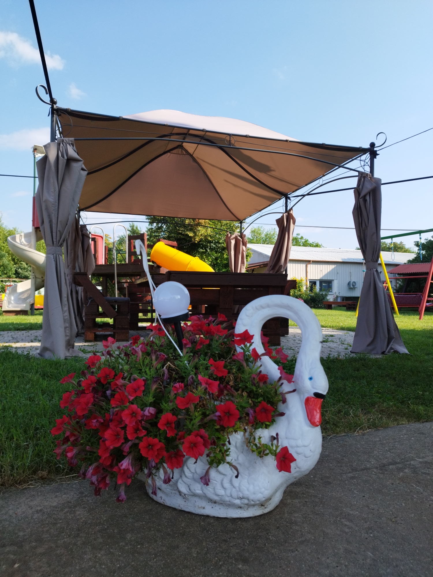 The image shows a bright outdoor scene, likely in a public space like a park or backyard, on a clear day. In the foreground, a white planter shaped like a swan is filled with vibrant red petunias. The swan planter has blue eyes and a red beak, and a small solar-powered light is positioned in front of it. Behind the planter, a brown canopy tent with draped grey curtains provides shade over a wooden picnic table and benches. In the background, there is playground equipment, including a yellow slide and a swing set with red accents. A low-lying building with a white metal roof is visible further in the background, along with trees and a clear blue sky. The overall impression is of a well-maintained recreational area. The image does not contain any individuals. No specific location in Bucharest, Romania can be identified from the visual information provided.