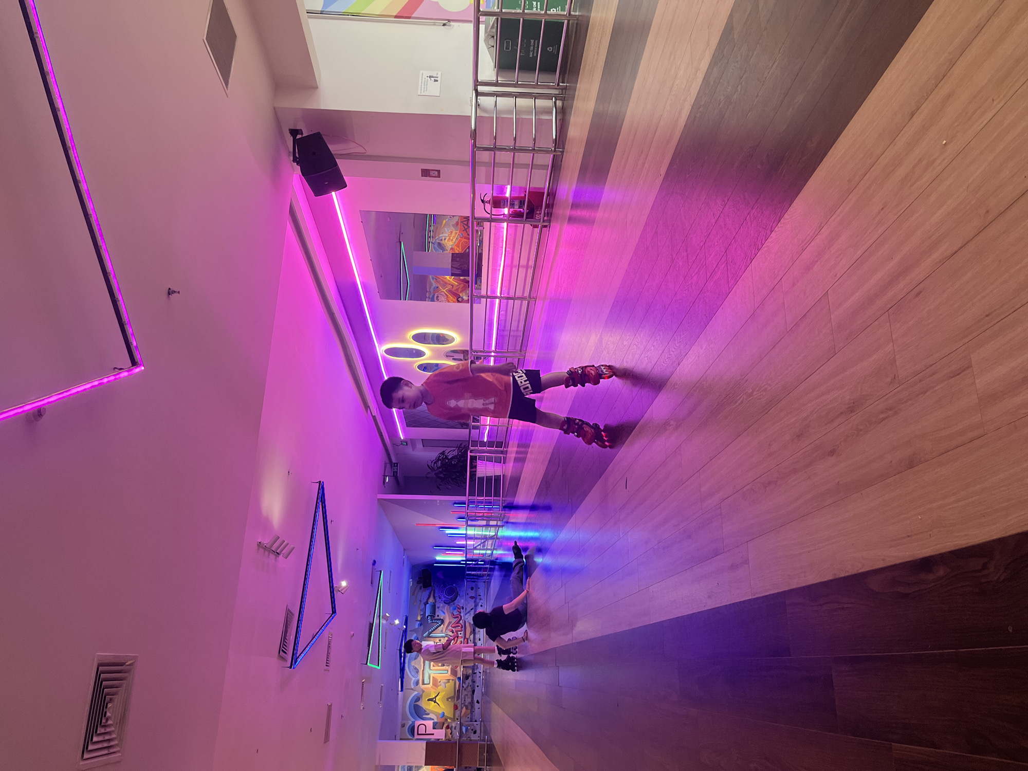 A brightly lit roller skating rink with two children visible. The rink has alternating brown and light-colored wood planks. The walls are adorned with neon lights of various shapes and colors, including pink, blue, and green. One child is skating closer to the camera while the other is further down the rink. A metal railing is visible along one side of the rink. A speaker is mounted on the wall near one of the neon light fixtures. A graffiti-style design can be seen on one wall further down the rink.