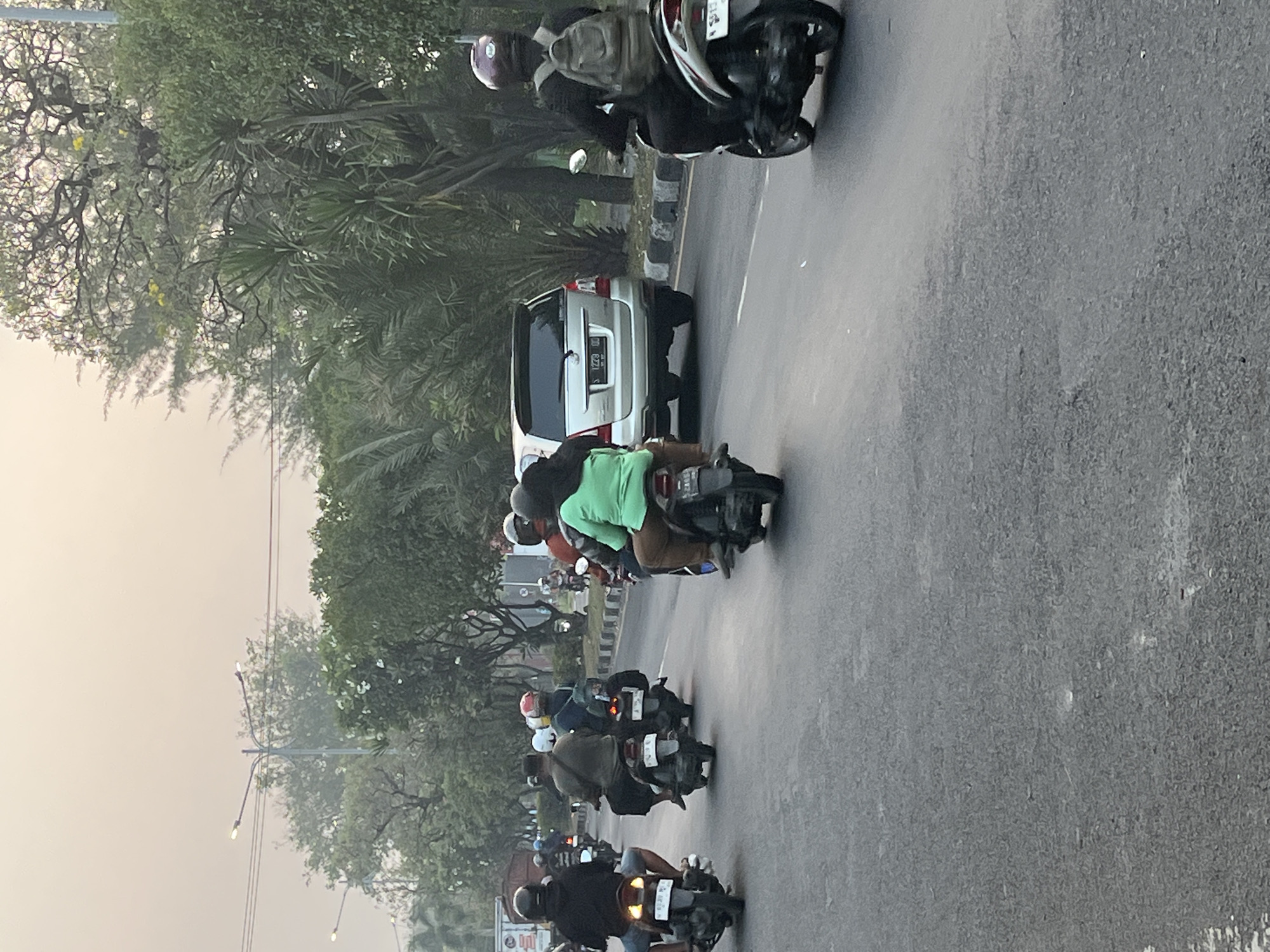 The image depicts a street scene in Kludan, Indonesia, likely during the late afternoon or early evening, given the soft, diffused lighting. A group of people are traveling on motorbikes along an asphalt road. In the foreground, a motorbike with two occupants is visible, with the rider wearing a helmet and the passenger wearing a green shirt. To the left, several other motorbikes with riders and passengers, all wearing helmets, are also traveling in the same direction. A silver SUV is in the middle ground, to the right of the motorbikes. Trees and foliage line the side of the road, and streetlights are visible in the background. The overall atmosphere suggests everyday life and transportation in a tropical or subtropical urban or suburban environment.