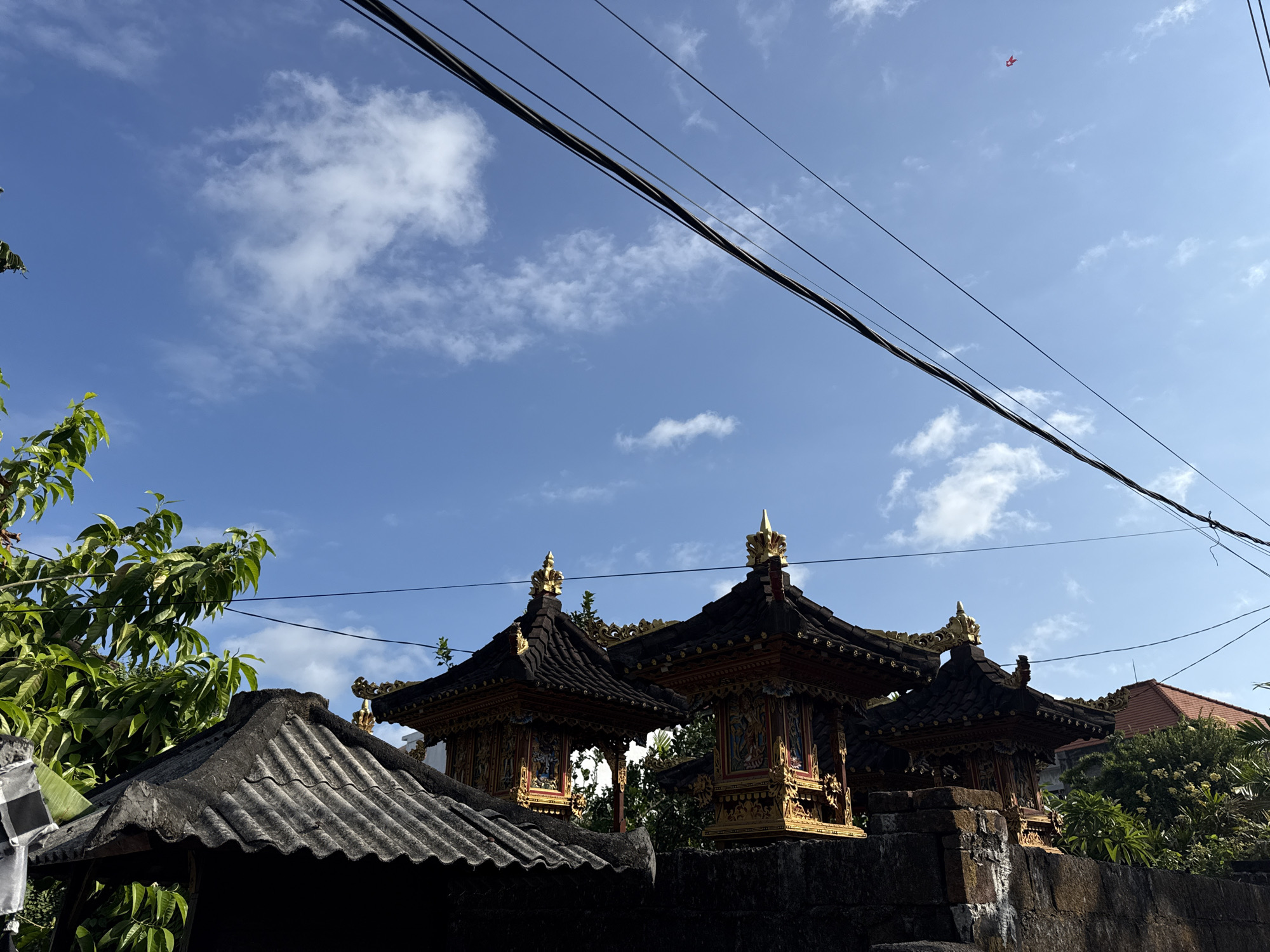 The image depicts an outdoor scene with traditional Balinese structures against a bright blue sky with scattered white clouds. In the foreground, a dark, textured roof of a smaller structure is visible, partially obscured by lush green foliage to the left. Behind this, three elaborately decorated, multi-tiered structures with dark tiled roofs and ornate golden detailing stand on a stone wall. These appear to be shrines or temples, common architectural elements in Bali. Overhead, thick black electrical wires stretch across the frame. In the distance, a tiny red object, possibly a kite, is visible in the sky. The overall environment suggests a tropical or subtropical location, consistent with Denpasar, Indonesia, known for its rich cultural heritage and architecture. The lighting indicates it is daytime, likely morning or afternoon, with clear weather. No people are present in the frame.
