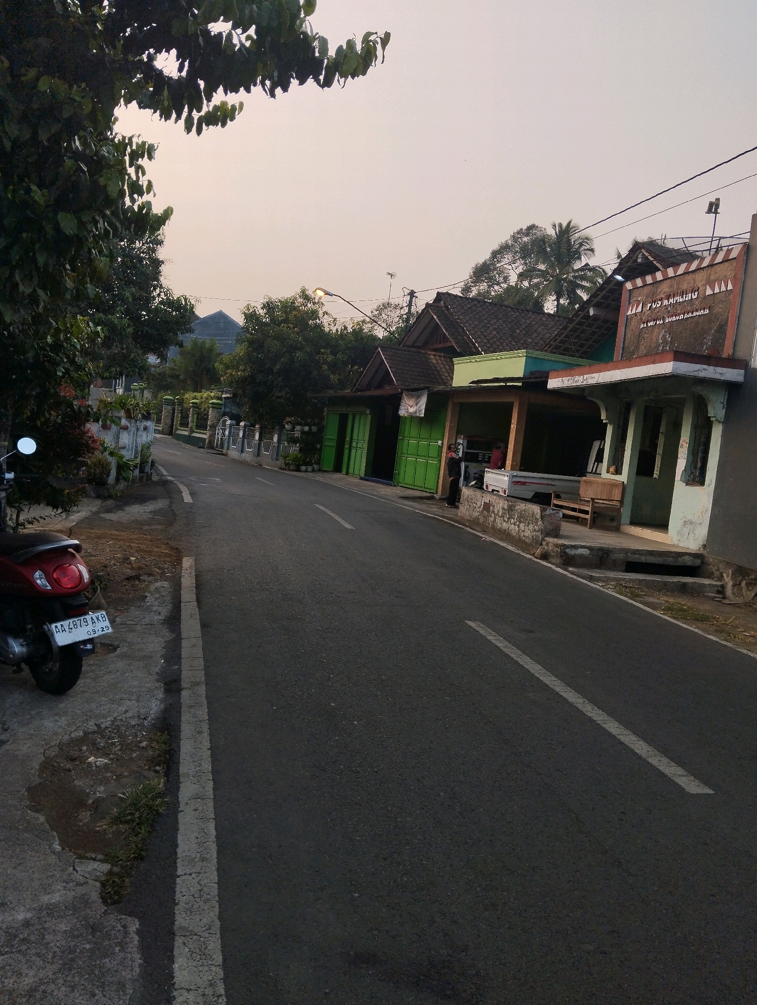 The image depicts a street scene in Salatiga, Indonesia, likely in a rural or suburban area given the presence of trees and low-rise buildings. The time of day appears to be late afternoon or early evening, as indicated by the soft, diffused light and the pale sky.
On the left side of the road, a red scooter is parked on the side. Its license plate reads "AA 4879 AKB 09-29". Adjacent to the scooter, there's a concrete walkway with some patchy vegetation and a small puddle of water. A mature tree with dense foliage overhangs the walkway and the road.
The road itself is asphalt, with a white dashed line marking the center. On the right side of the road, a row of modest buildings with pitched roofs are visible. These appear to be small shops or houses, some with open-fronted areas. One building has a sign with text that is partially legible, appearing to say "TUS KAMLING" and below it, "AL UYUT GUNUNG KRAJAN". A white pickup truck is parked in front of one of the shops, and a person is standing near it. Another person is visible standing in the doorway of a building with green doors. There are potted plants and decorative elements along the buildings and in front of them.
Further down the street, more buildings and trees are visible, along with a street lamp. The overall impression is of a quiet, everyday street in a Indonesian town.