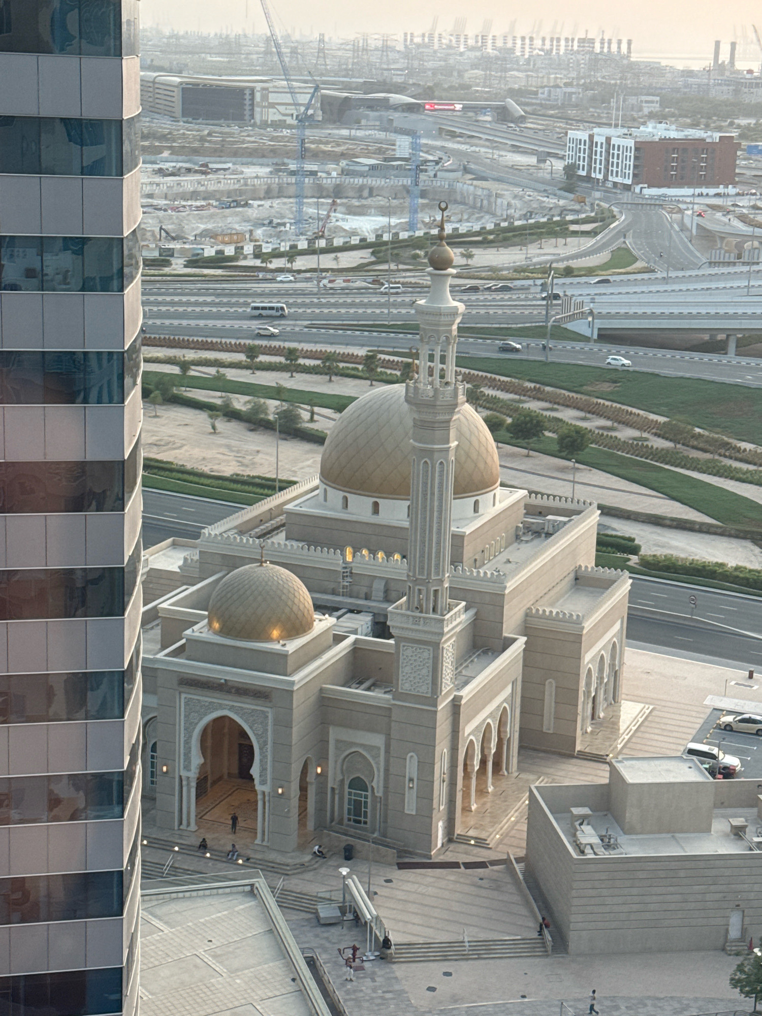 The image depicts a bird's-eye view of a mosque in Dubai, United Arab Emirates, with a modern skyscraper in the foreground on the left. The mosque features golden domes and a tall minaret, constructed from beige stone with arched windows and entrances. Several people are visible in and around the mosque, including some sitting on steps outside. In the background, a busy highway overpass system with cars and buses is visible, along with ongoing construction sites with cranes and infrastructure development. The distant skyline shows industrial elements and buildings under a hazy sky.