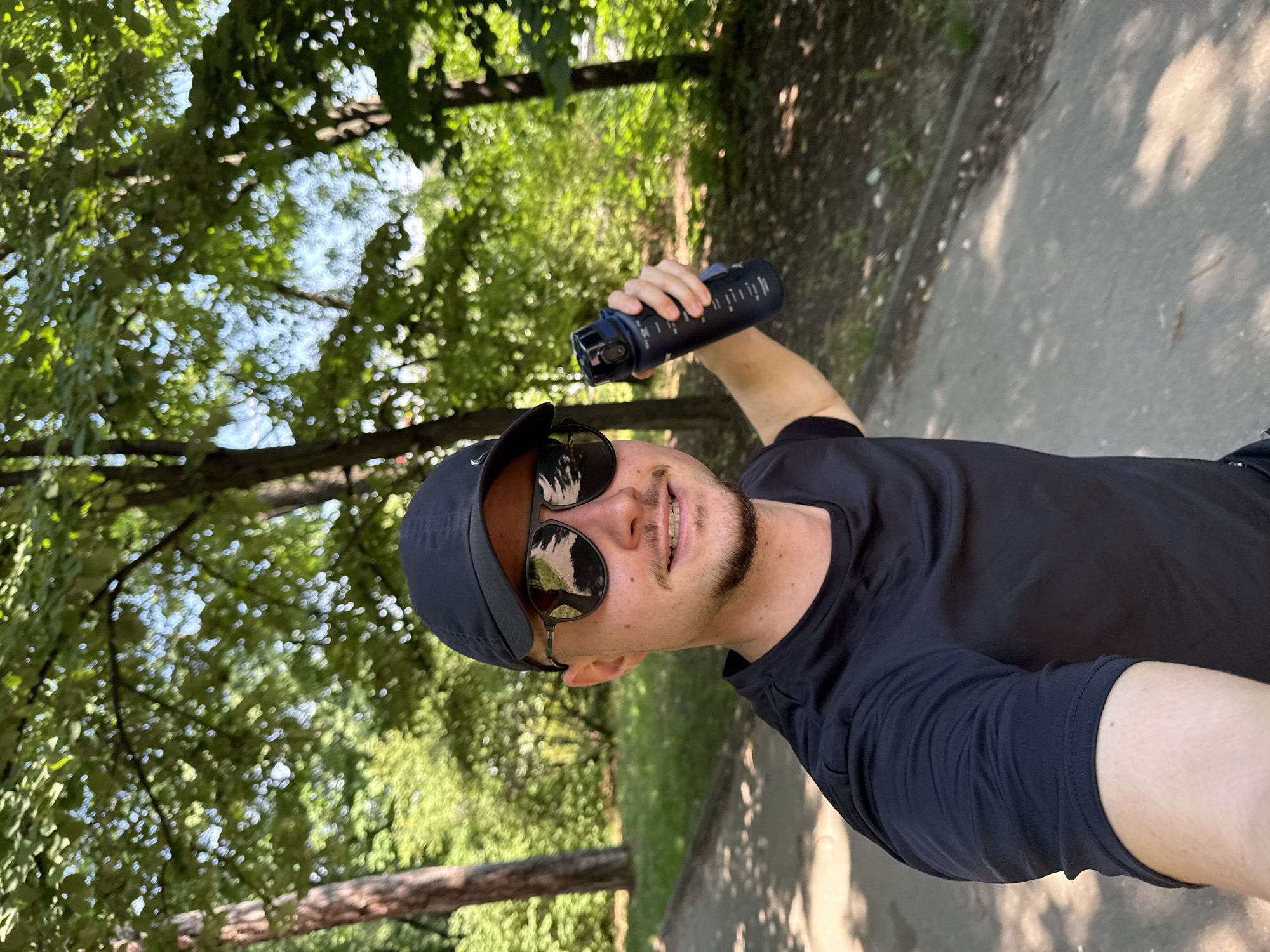A man wearing a black t-shirt and a black baseball cap is taking a selfie outdoors. He is also wearing sunglasses and has a slight beard and mustache. In his right hand, he is holding a dark-colored water bottle. The man is smiling at the camera. The background shows a park-like setting with trees and a paved path. The scene appears to be in Sofia, Bulgaria, as suggested by the presence of lush greenery and mature trees common in urban parks within the city. The lighting indicates it is daytime, likely sunny, casting shadows from the trees onto the path.