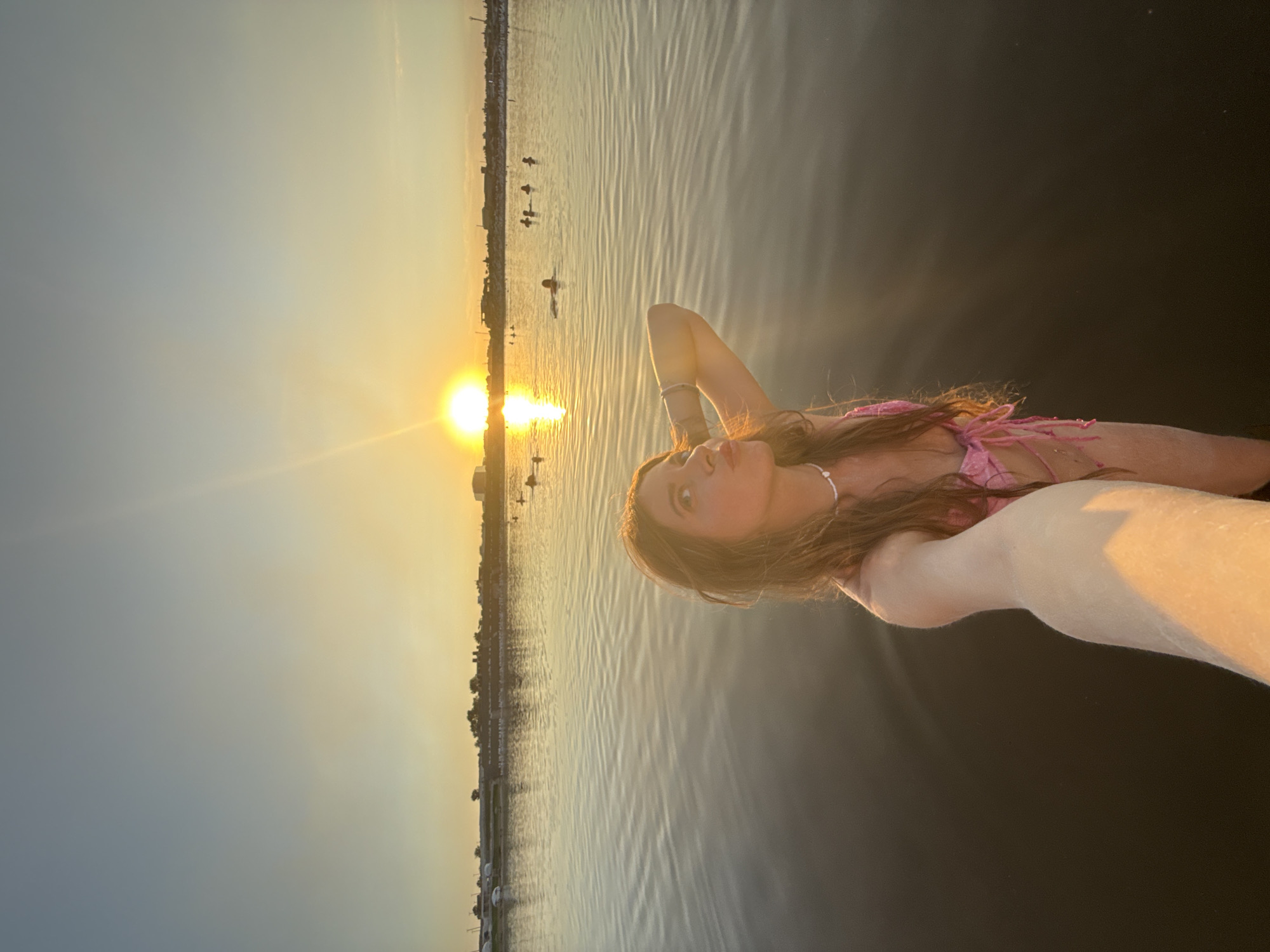 A young woman in a pink bikini stands in shallow water, holding a camera out to take a selfie. She is facing the camera with her head tilted back and her lips pursed. Her right arm is bent at the elbow, with her hand resting on the back of her head. The sun is setting on the horizon behind her, casting a warm glow across the water and sky. The water is calm, with gentle ripples reflecting the light. In the background, a city skyline is visible, with buildings and trees lining the shore. Several other people are in the water in the distance, swimming and enjoying the sunset. The image appears to have been taken in Sol-Iletsk, Russia, known for its saline lakes and resorts.