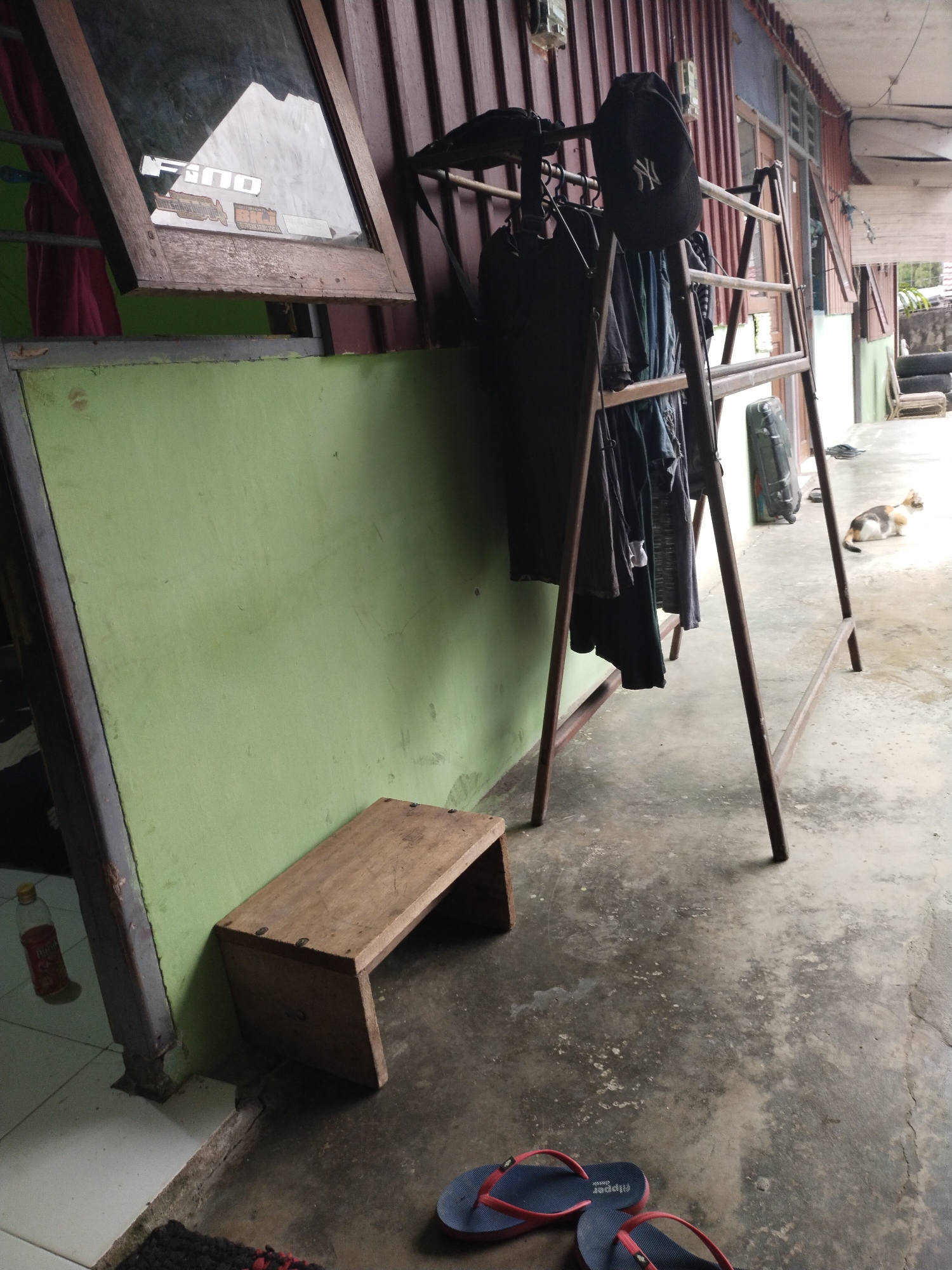 The image depicts an outdoor scene, likely a covered walkway or porch area adjacent to a building with a corrugated metal roof and wooden walls. In the foreground, two pairs of flip-flops are on the concrete ground. To the left, a green wall is partially visible, with a wooden stool in front of it. Further back, a metal clothes drying rack is laden with dark-colored clothing, including a baseball cap with a white logo. A dark suitcase is propped up near the drying rack. In the background, a calico cat is resting on the concrete floor. The overall environment appears to be a residential or utilitarian outdoor space. The presence of the building materials and the general upkeep suggest a location in Manokwari Barat, Indonesia.