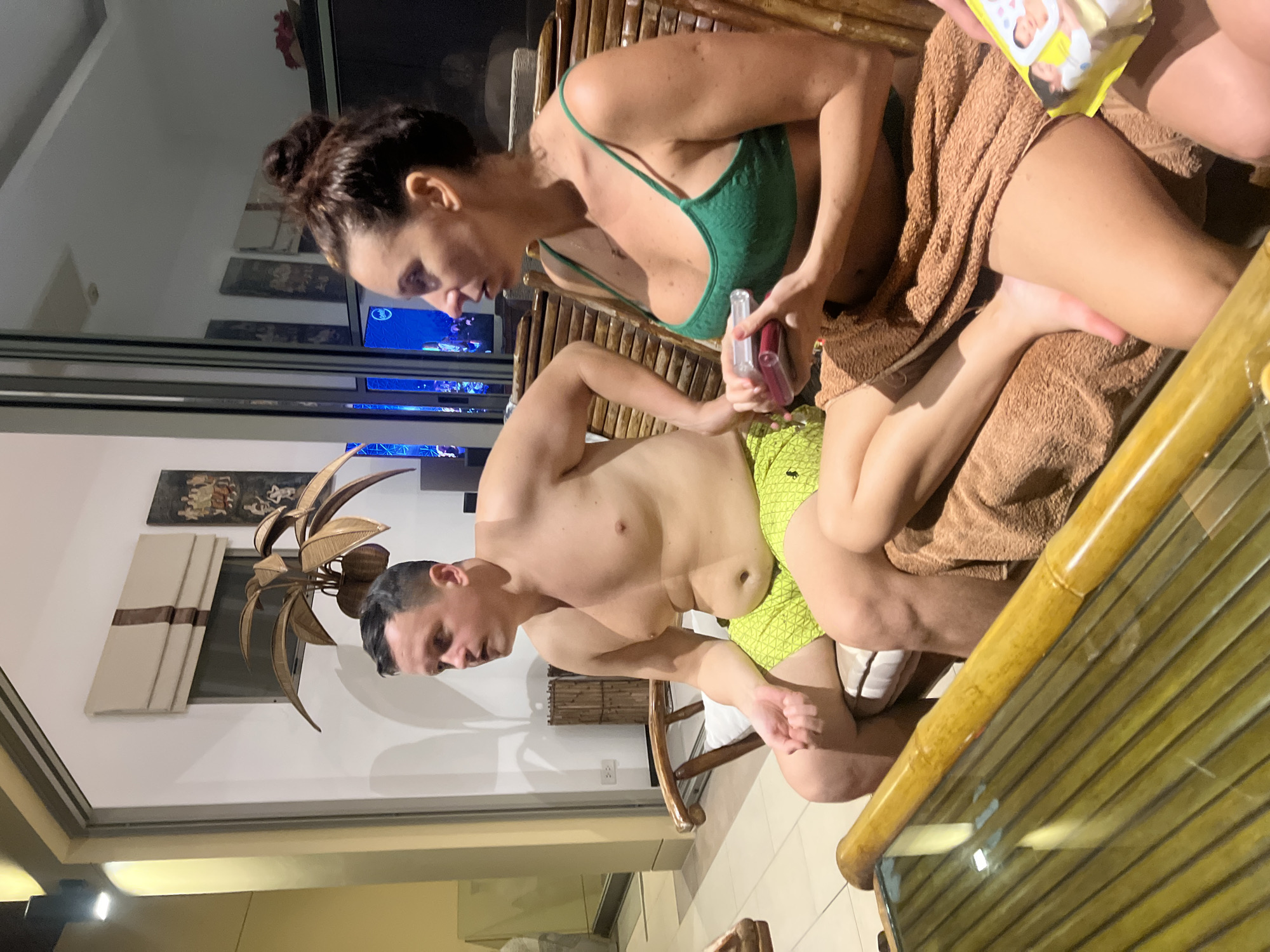 A man and a woman are seated on bamboo furniture in what appears to be an outdoor or semi-outdoor living area, likely in Baan Bo Phut, Thailand. The man, to the left, is wearing lime green swim trunks and is shirtless. The woman, to the right, is wearing a green bikini top and a brown towel wrapped around her lower body. She is holding two small containers, one clear and one red. The furniture is made of bamboo, and a bamboo coffee table with a glass top is in the foreground. In the background, there are large glass doors, artwork on the wall, and a tropical plant. The lighting suggests it is evening or night.