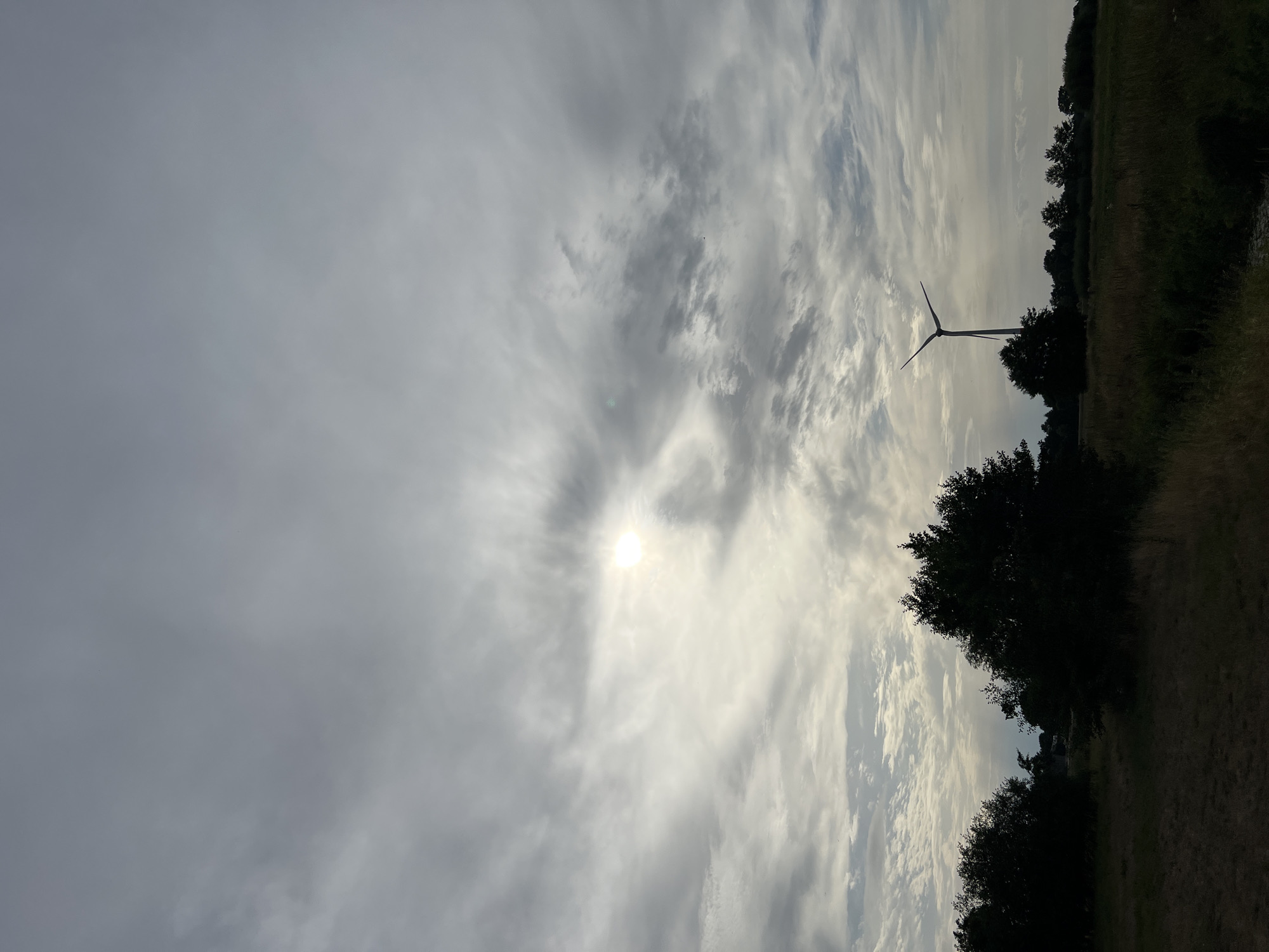 A solitary wind turbine stands silhouetted against a cloudy sky. The sun, partially obscured by clouds, casts a faint glow. The foreground is a dark, grassy field with scattered trees. The overall impression is one of a rural landscape on an overcast day. The location is identified as Unknown, Unknown.