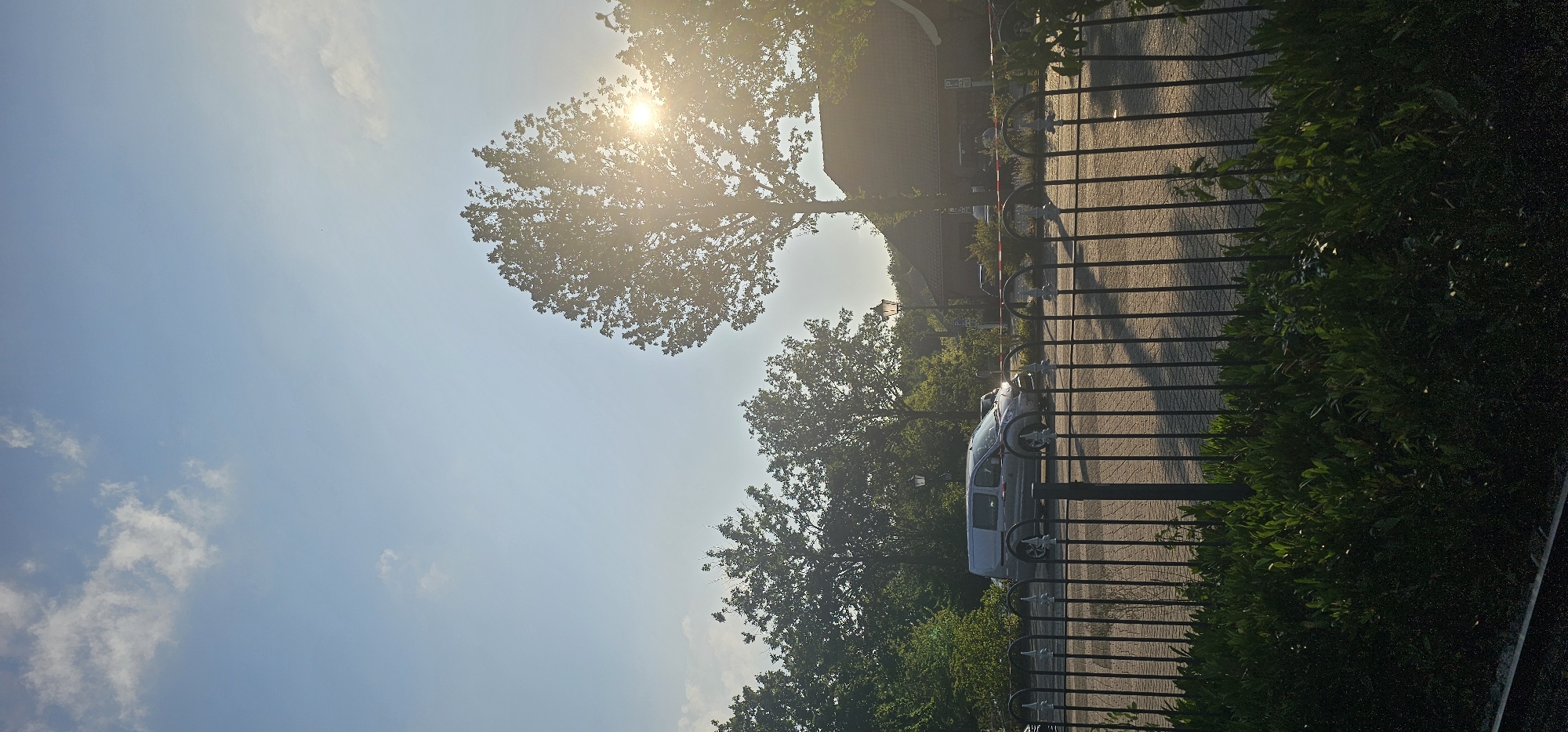 The image depicts a sun-drenched scene on a paved road, likely in Groenlo, Netherlands, with a black metal fence in the foreground. A silver van is driving on the road, casting a shadow. Beyond the fence, lush green foliage and trees are visible, with a large tree to the right of the frame partially obscuring the bright sun, which creates lens flare. In the background, a building with a dark roof and brickwork is visible, along with a lamppost and other trees. The sky is blue with scattered clouds.