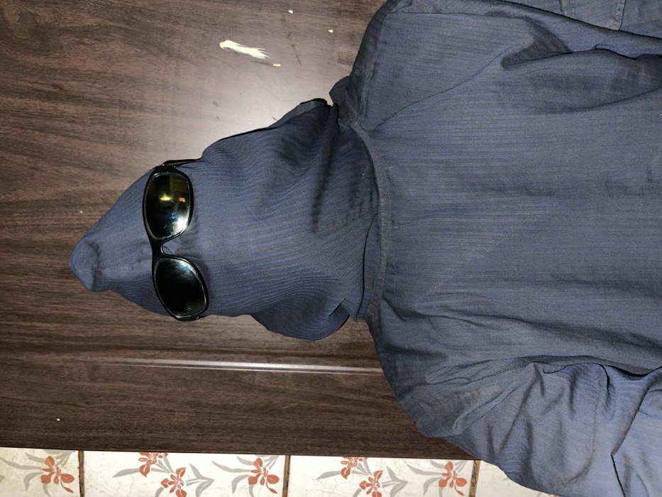 A dark blue button-down shirt lies on a surface transitioning from brown wood to tiled floral patterns. The shirt's left sleeve is bundled, concealing a head-like shape. Dark sunglasses sit on this form, mimicking eyes. A small dab of white is on the wood surface above the "head". The shirt's fabric has subtle, fine lines, adding texture to its appearance.