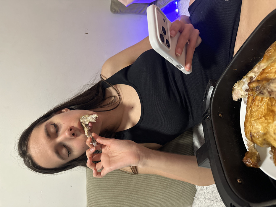 An indoor scene in Corby, United Kingdom, depicts a female individual eating cooked poultry. The subject, possessing long dark hair, wears a black ribbed tank top and is seated on a light-colored sofa or cushion. Her eyes are closed or cast downwards as she brings a bone, stripped of most meat but with some residue, to her mouth with her right hand. Her left hand holds a white smartphone. In the foreground, a black air fryer contains a white plate holding additional pieces of cooked, golden-brown poultry, some of which appear partially consumed. The background features a plain, light-colored wall, with blue string lights visible to the right.