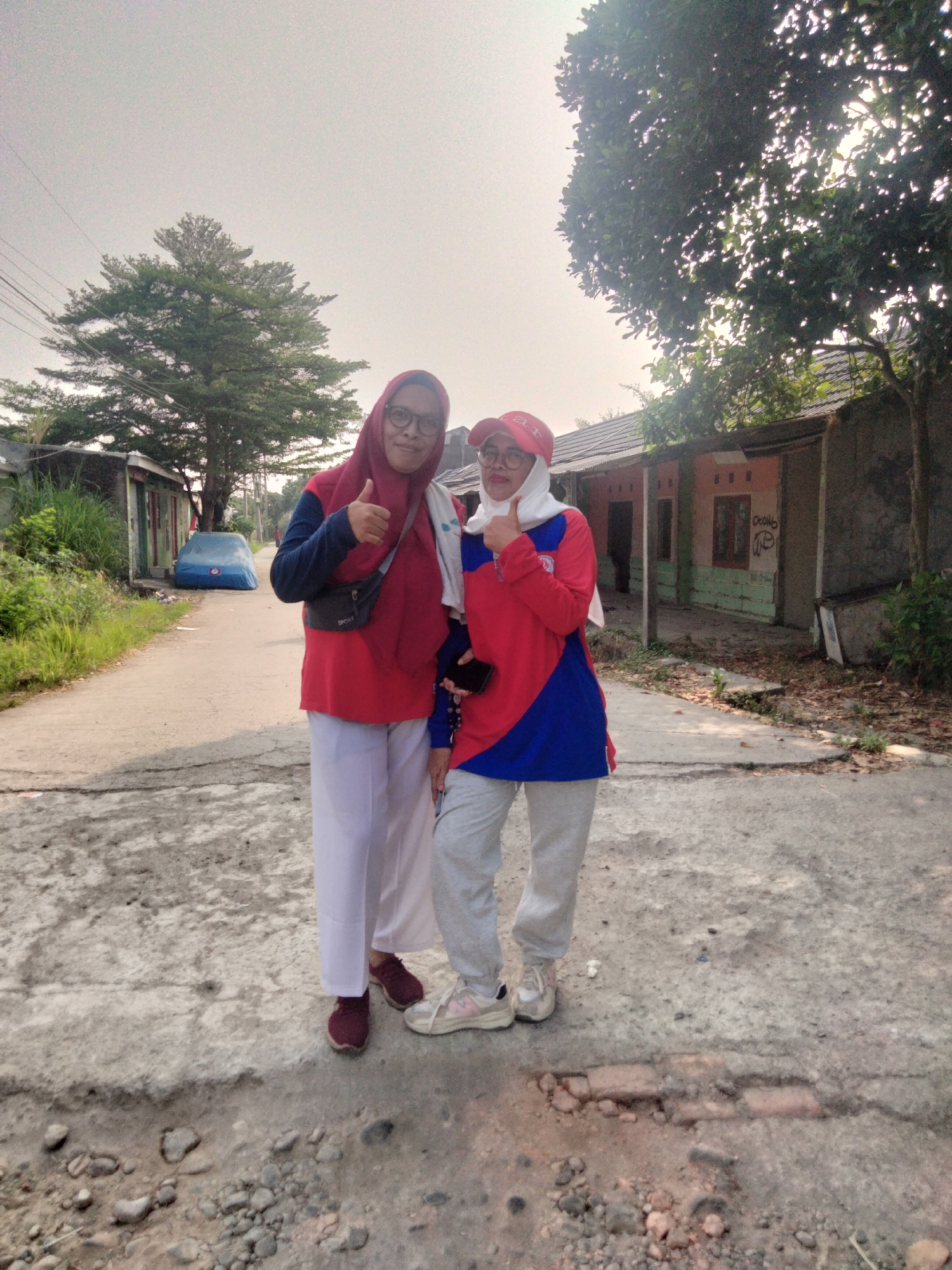 Two women stand on a paved road in Cariu, Indonesia. The woman on the left wears a red hijab, glasses, a red shirt with blue sleeves, and white pants. She gives a thumbs-up gesture. The woman on the right wears a white hijab and a red and blue shirt with gray sweatpants. She holds a black smartphone and also gives a thumbs-up. Both women are smiling. The road is lined with houses and vegetation. A blue tarp covers a car on the left side of the road. The sky is overcast.