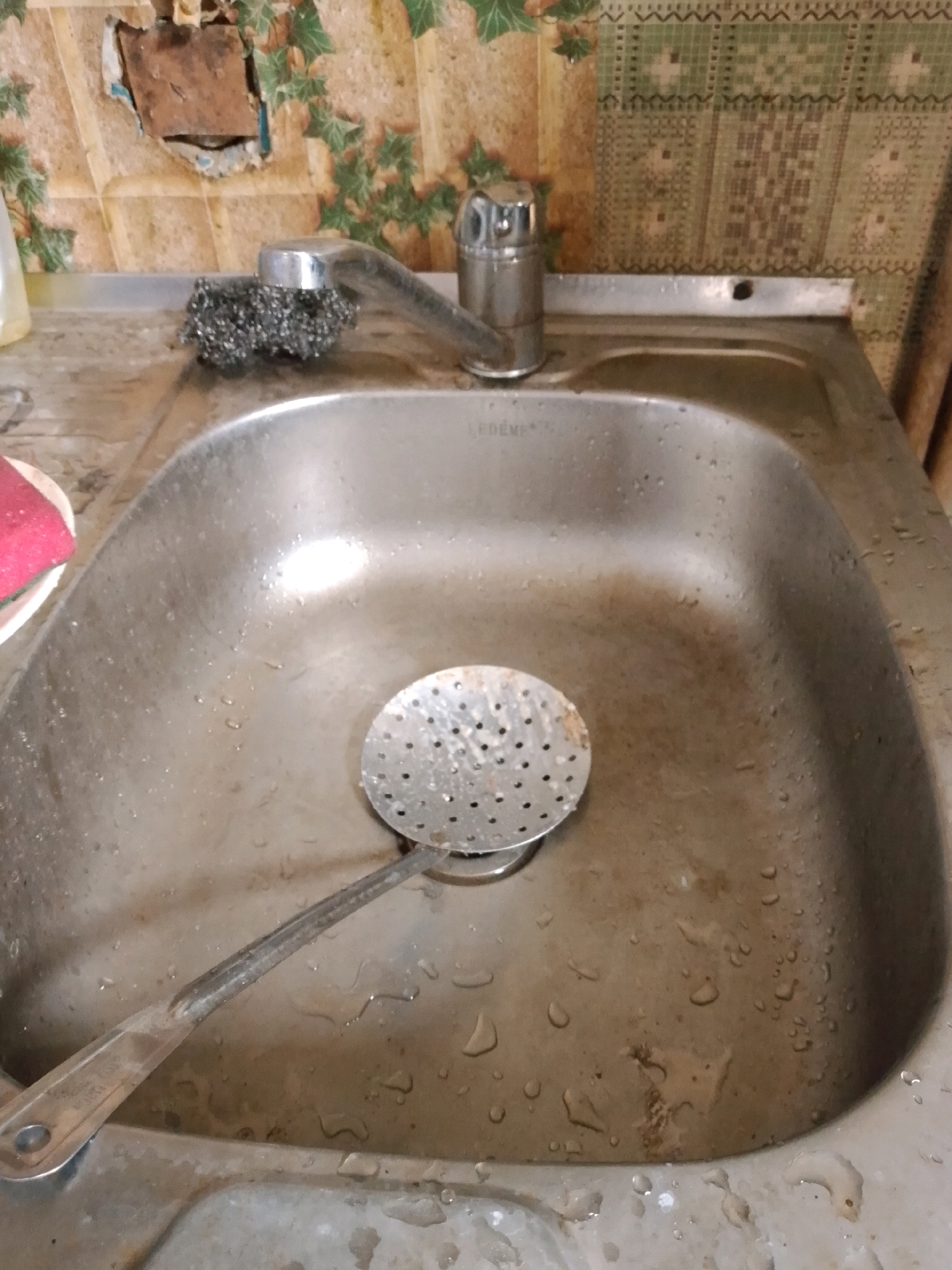 The image depicts a kitchen sink in what appears to be a utilitarian or possibly neglected environment. The sink itself is made of stainless steel and shows signs of wear and water spots. In the sink, there is a metal skimmer or slotted spoon. To the left, a scrub brush made of steel wool rests on the edge of the sink near the faucet. The backsplash behind the sink is covered in wallpaper with a floral pattern, but a section of it is torn away, revealing a rough surface underneath, possibly plaster or concrete. The faucet is made of chrome. The overall impression is one of a used and possibly unclean sink area. The location is identified as Ленінскі сельскі Савет, Belarus.